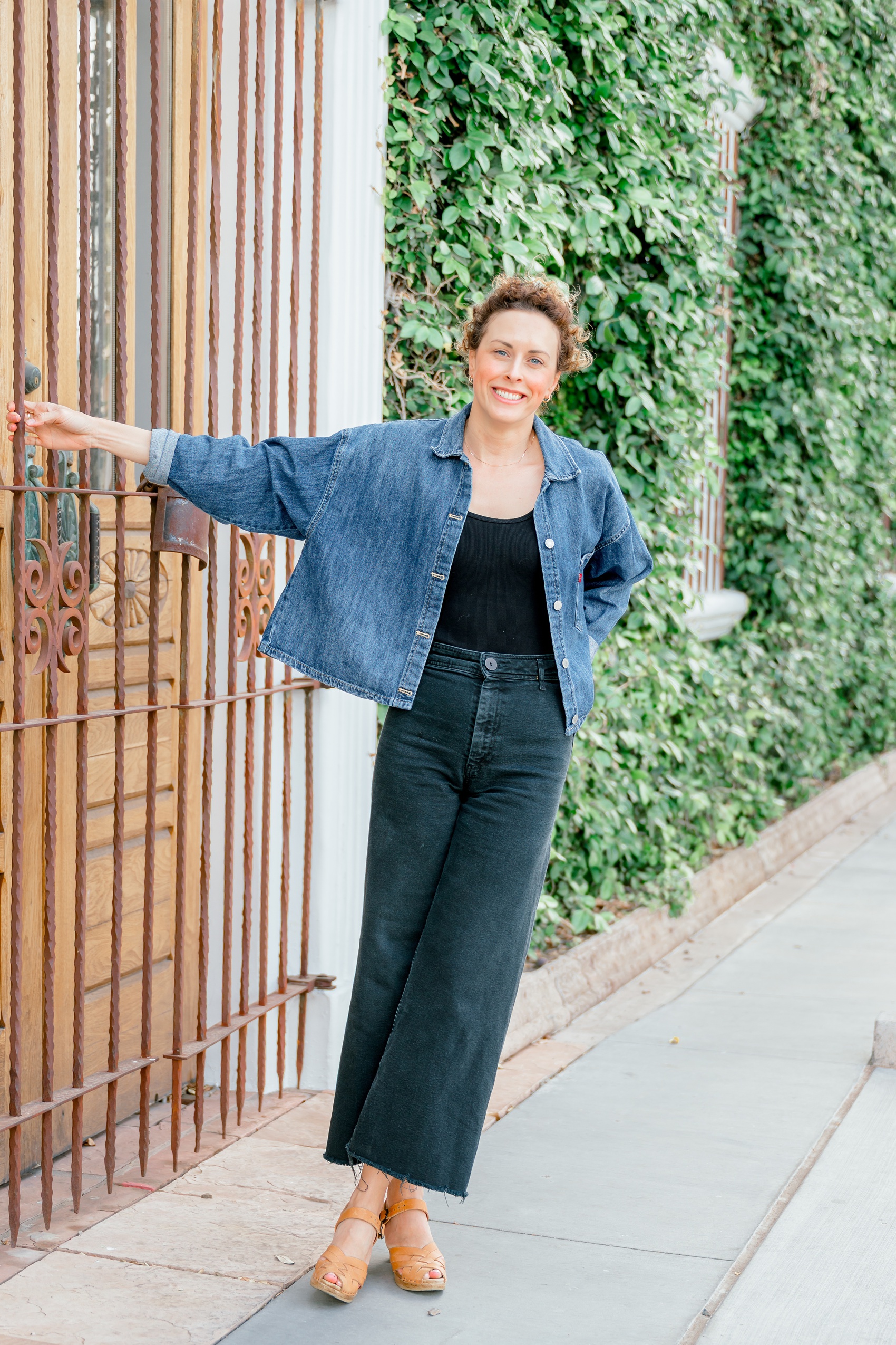 A woman leans while hanging on an iron gate to a building covered in vines in a denim jacket