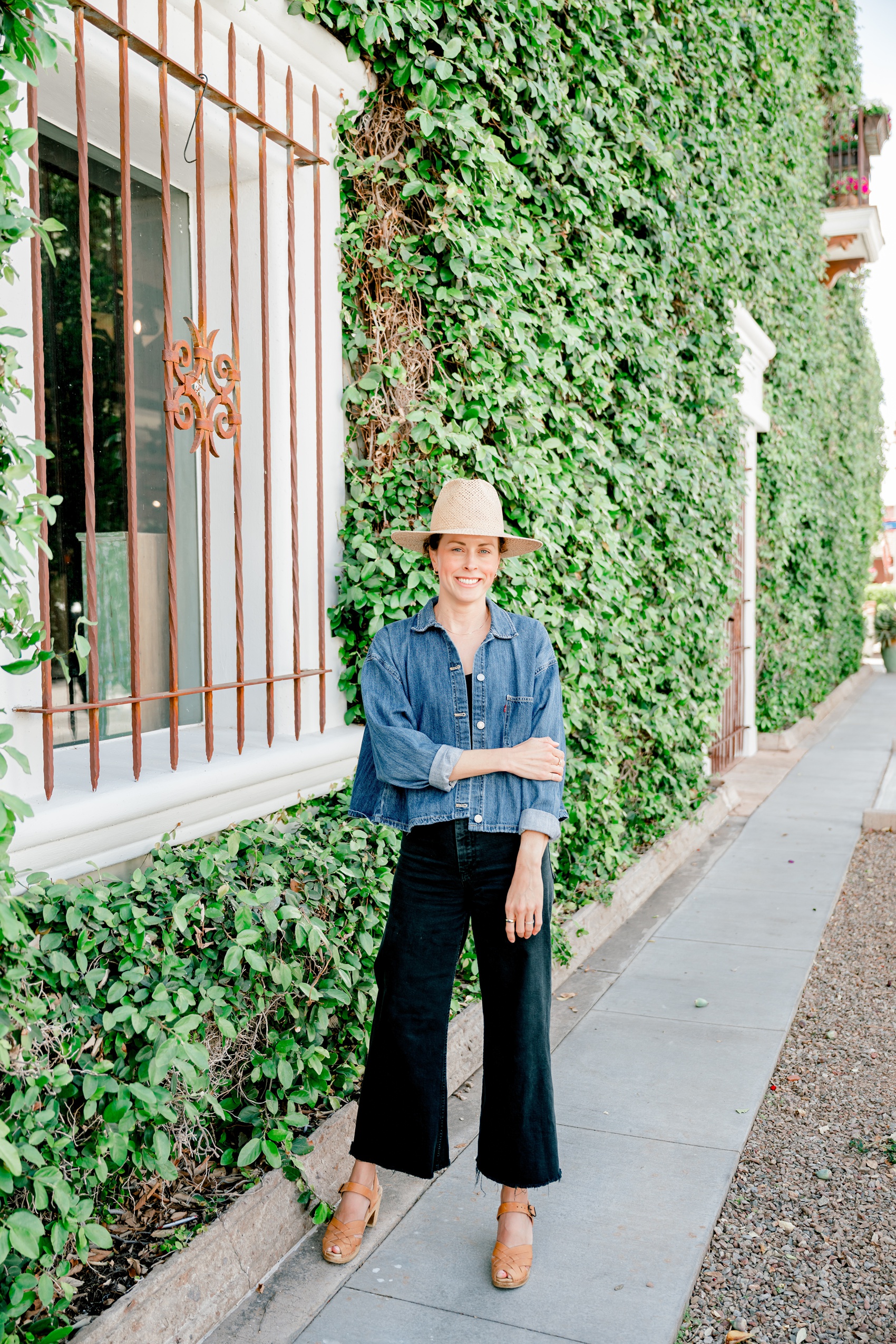 A woman holds her arm while standing on a sidewalk in front of a vine covered wall during her relaxed branding photos
