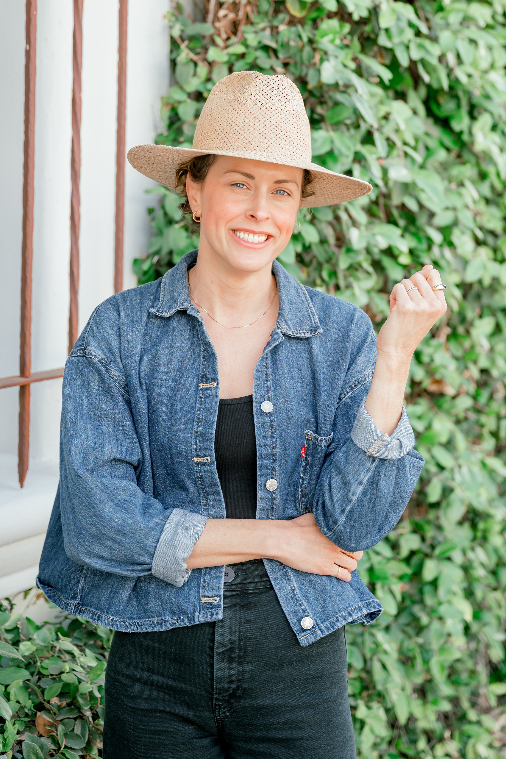 A woman in a denim jacket and woven hat smiles while standing in front of a vine covered wall for her relaxed branding photos