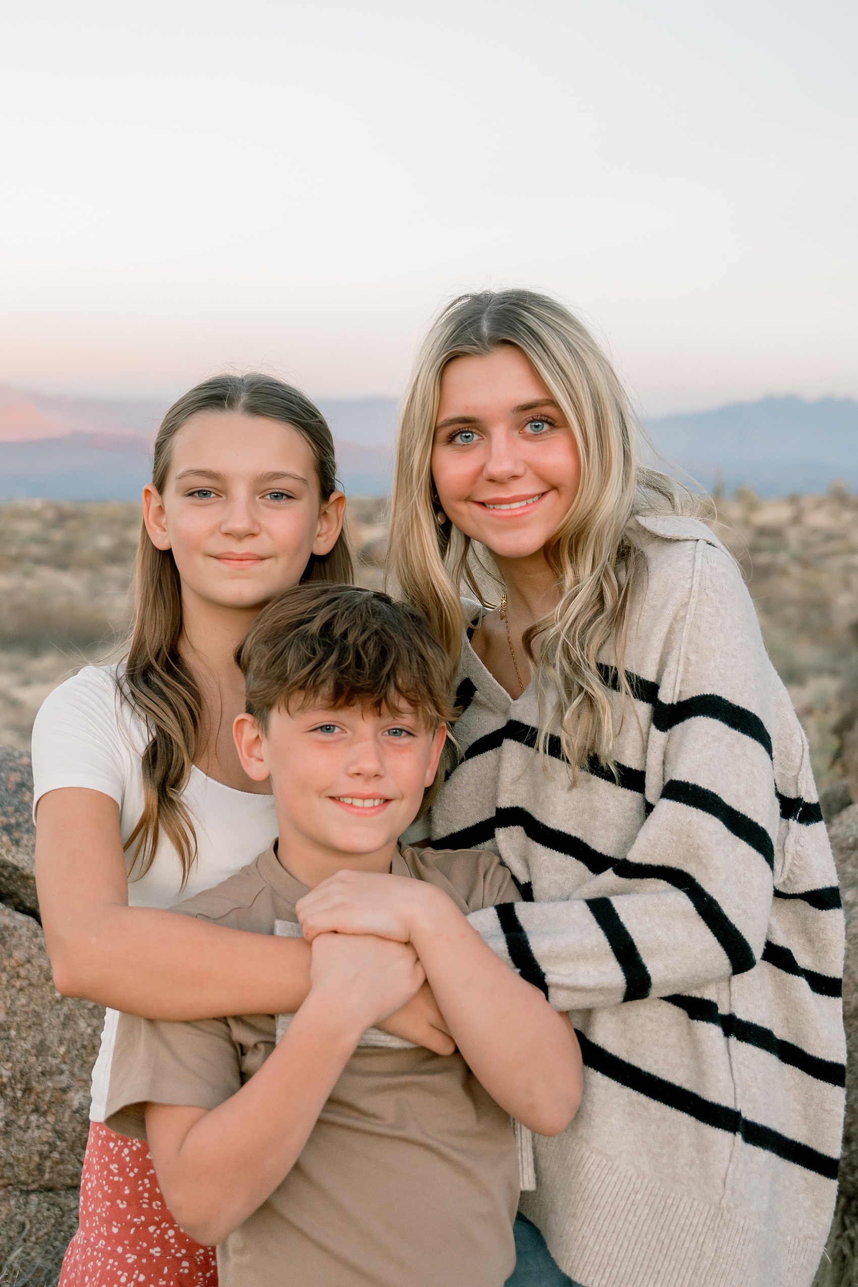 Three smiling siblings in neutral outfits hug at sunset on a desert trail during things to do in scottsdale with kids