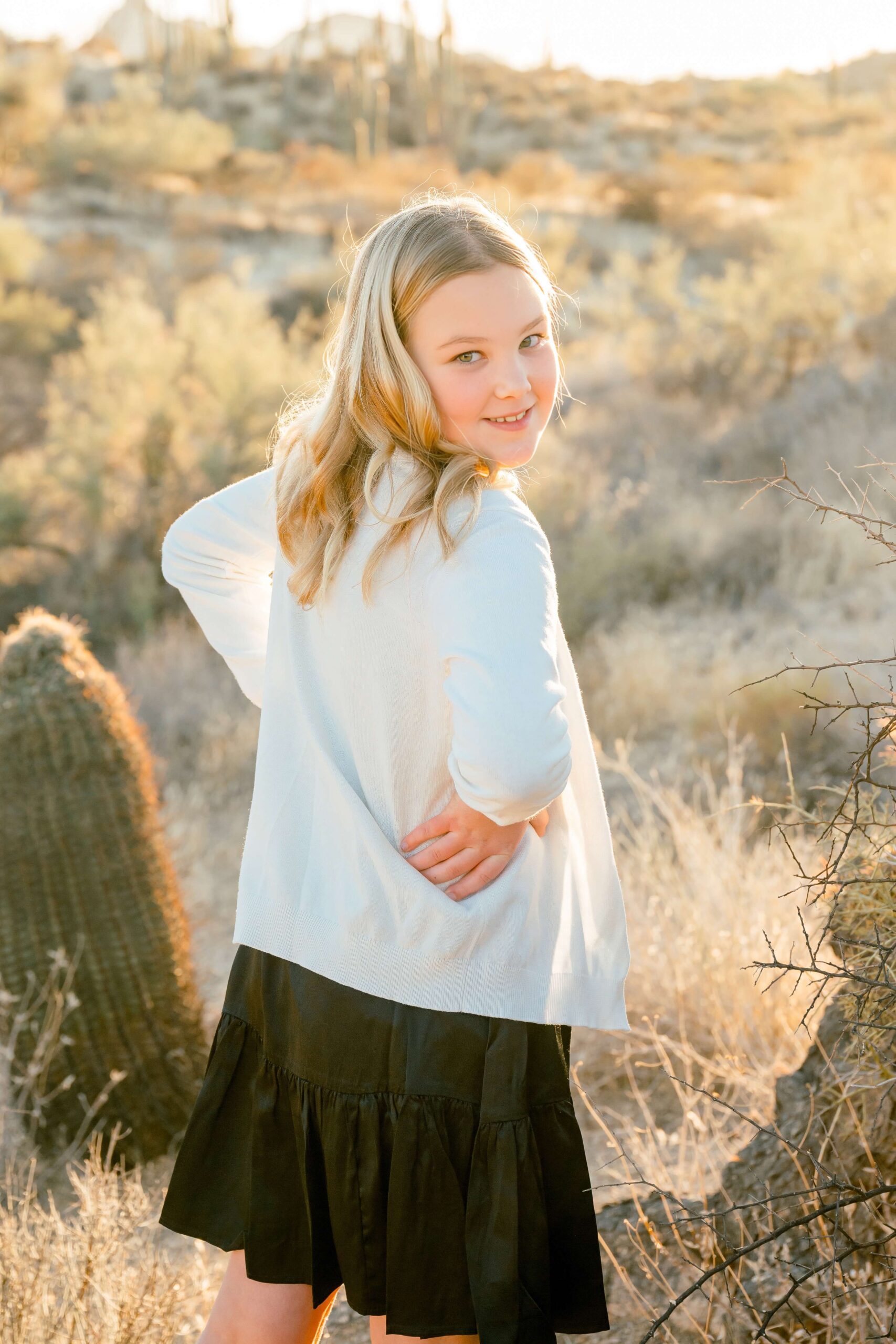 A young girl in a white sweater and black dress smiles over her shoulder while wlaking a trail in the desert at sunset