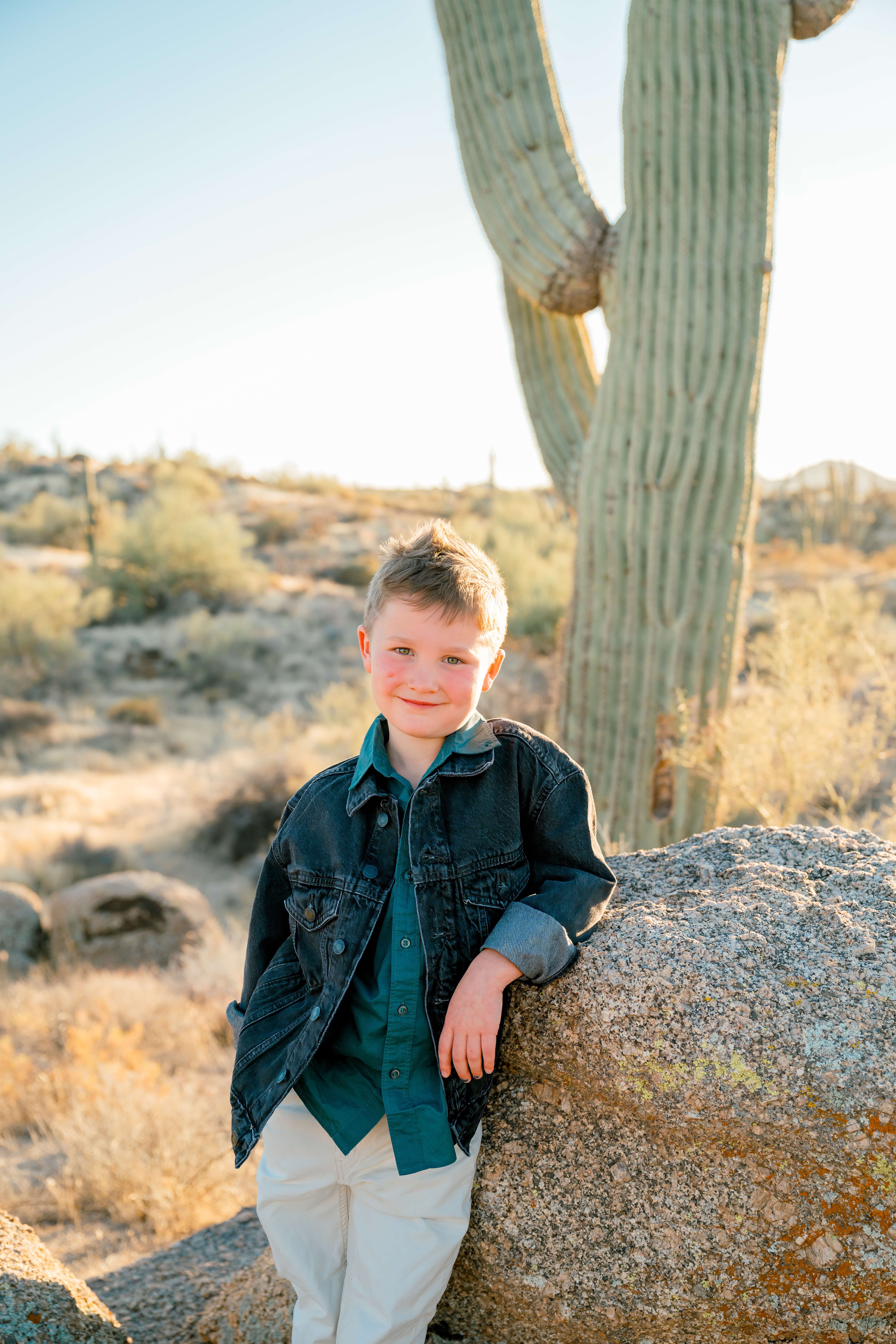 A young boy leans on a boulder in the desert in a denim jacket and khakis from children's boutiques in scottsdale