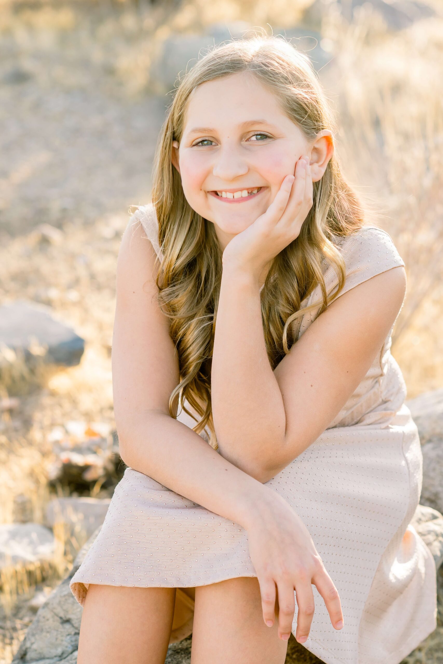 A young girl in a tan dress smiles while sitting on a rock at sunset after visiting children's boutiques in scottsdale