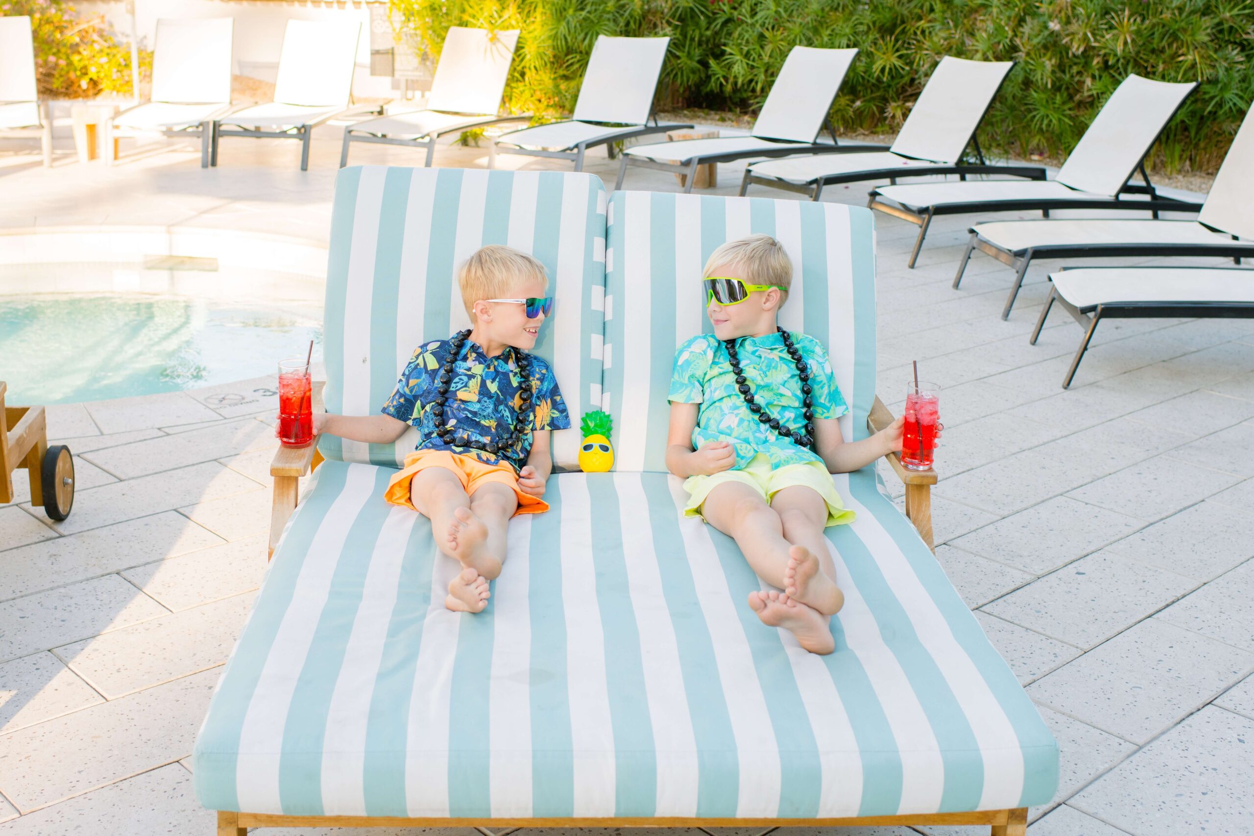 Two brothers in tropical shirts and sunglasses sip on red drinks on a large patio bed by the pool while visiting family friendly hotels in scottsdale