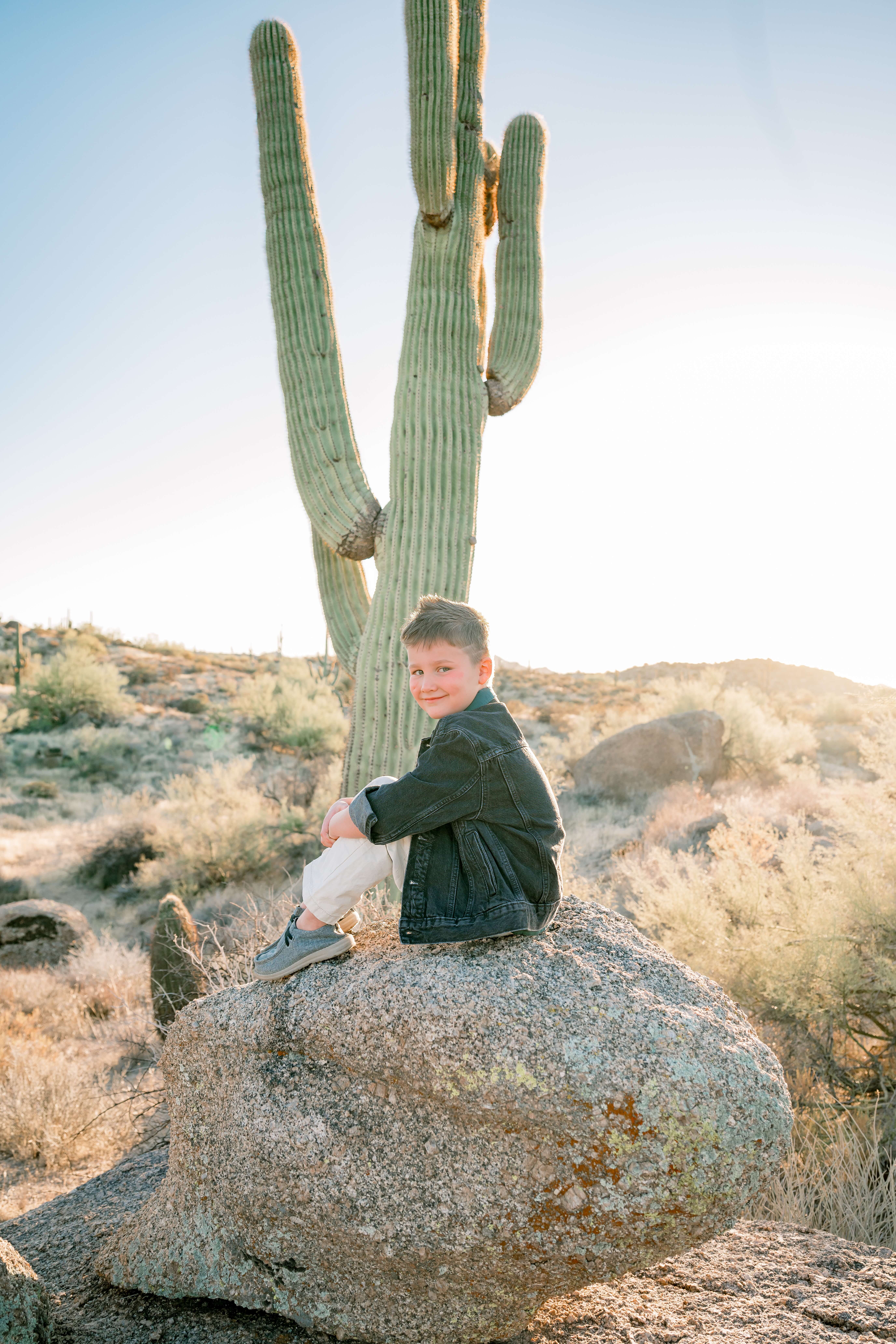 A young boy sits on a large boulder in the desert at sunset in white pants and denim jacket