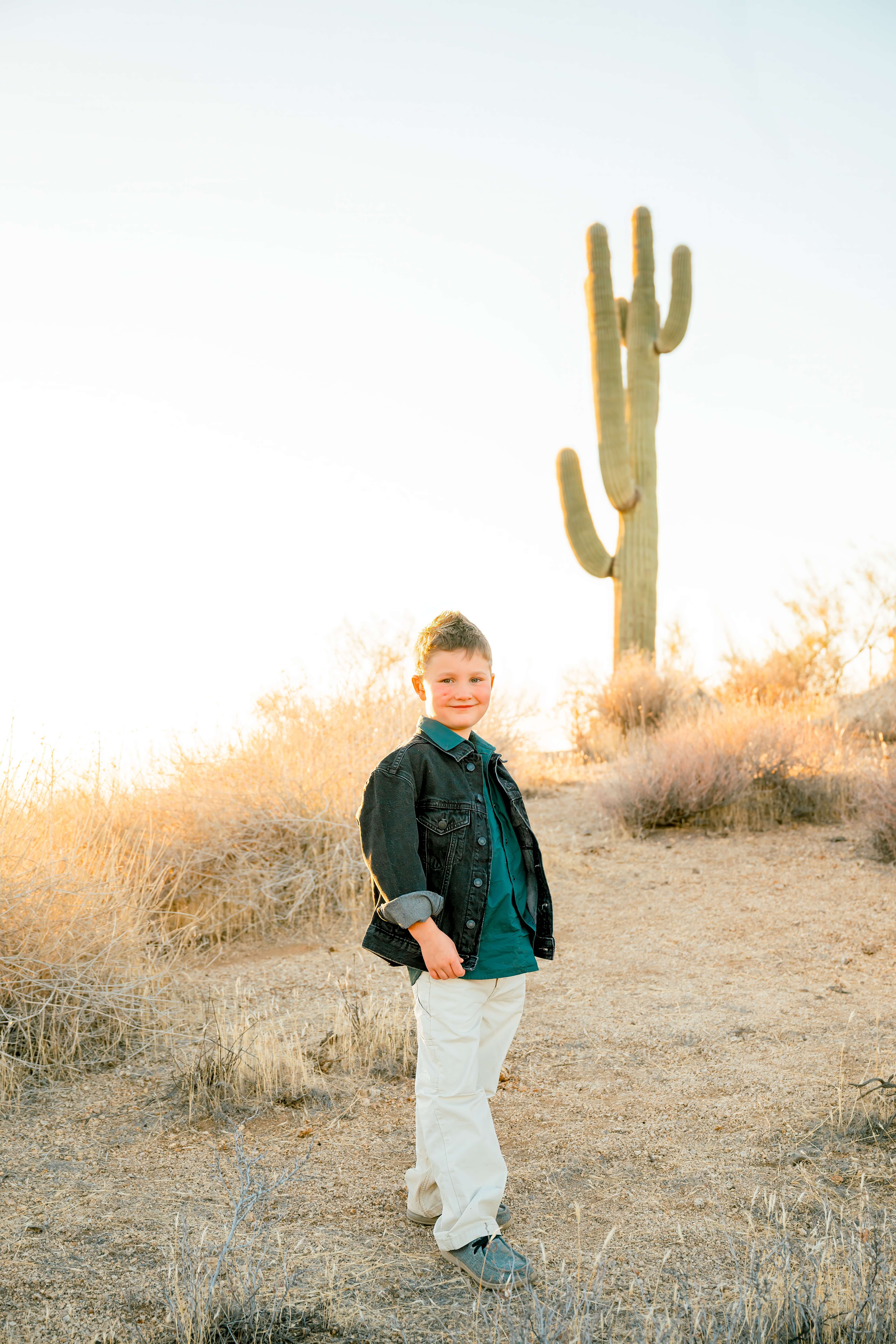A young boy in white pants and denim jacket stands in a trail at sunset smiling before visiting indoor playgrounds in phoenix