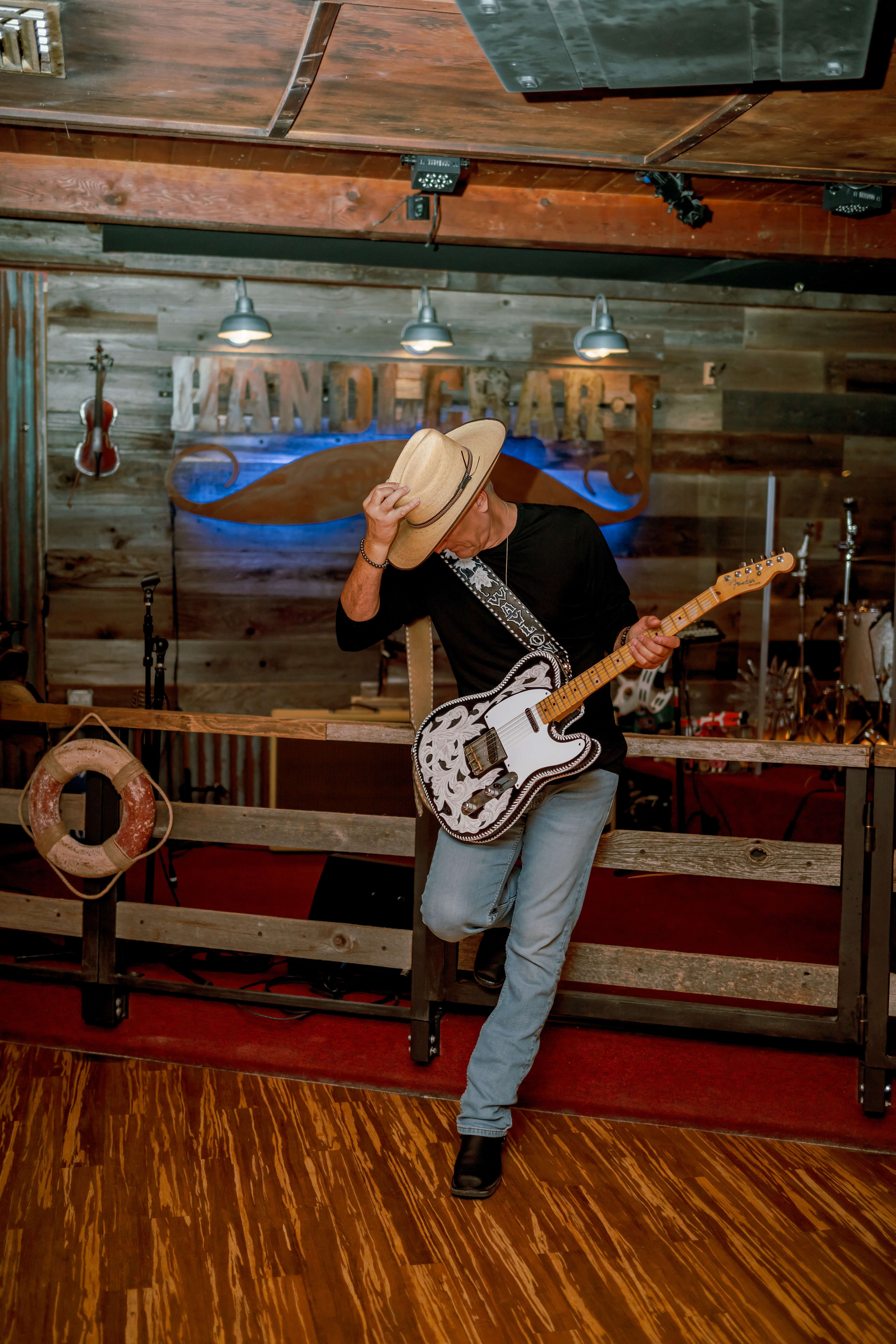 A man with a guitar leans on a wooden fence in a bar in jeans and cowboy hat