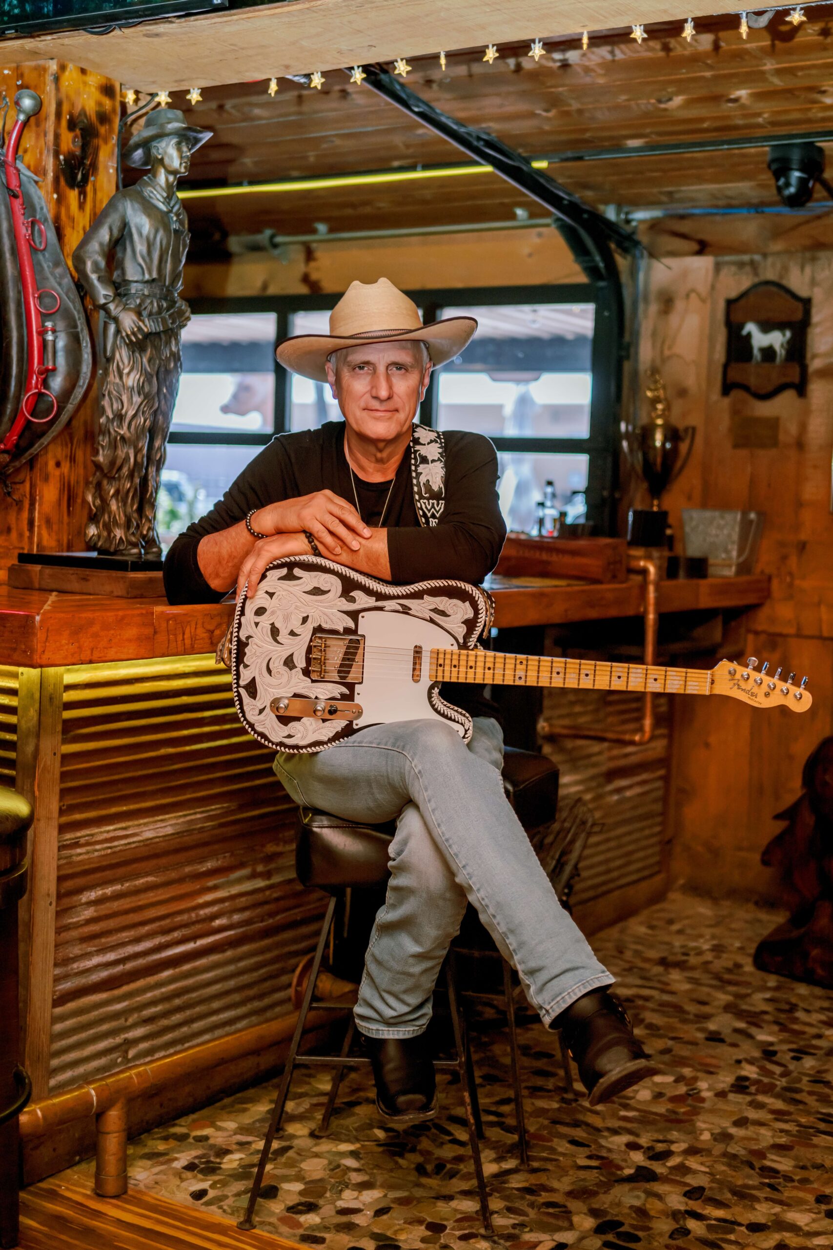 A guitar player with a black and white guitar in his lap sits on a bar stool in a western rustic bar