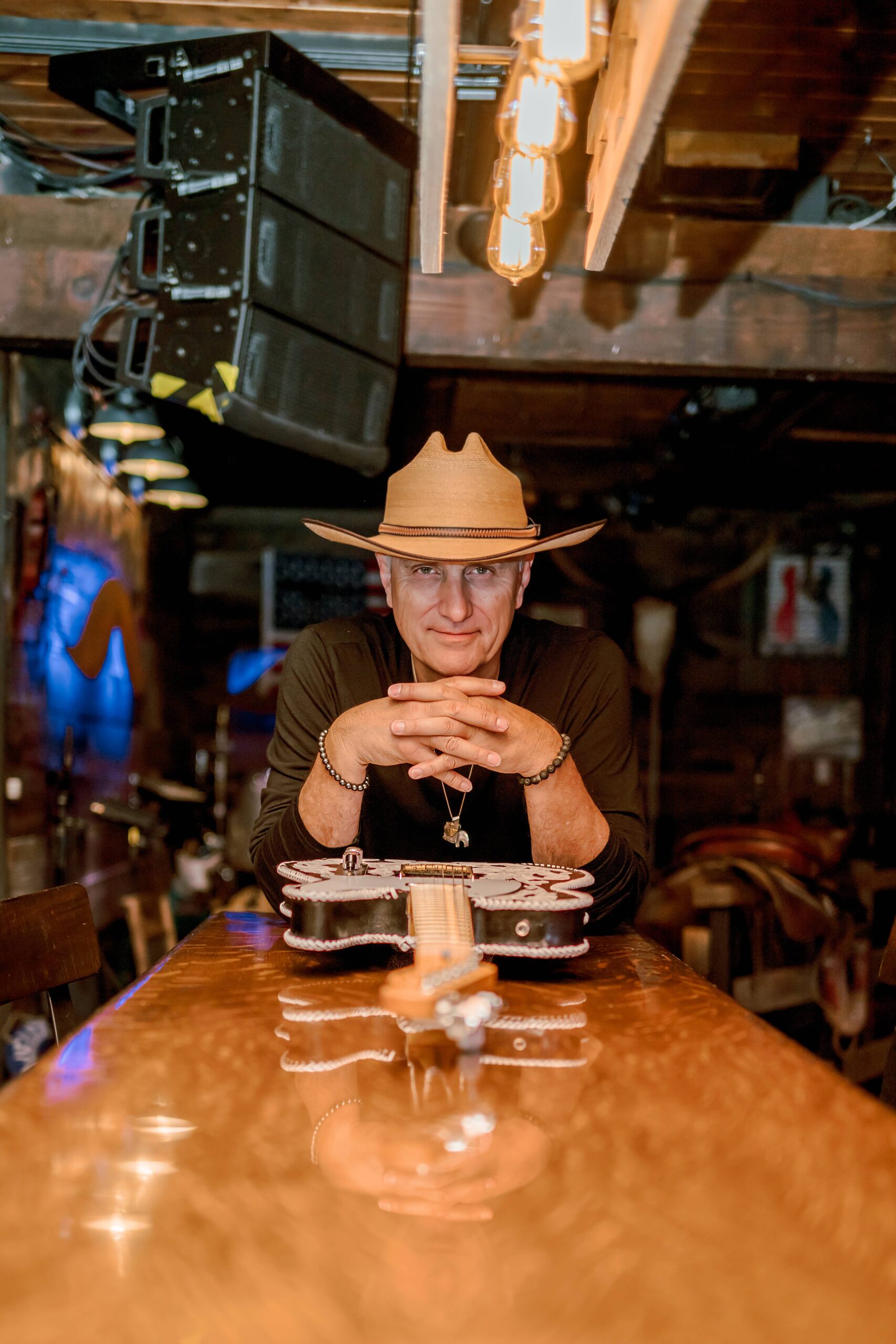 A man leans on a bar counter with a guitar laying on it in a balck shirt and tan cowboy hat during his personal branding session