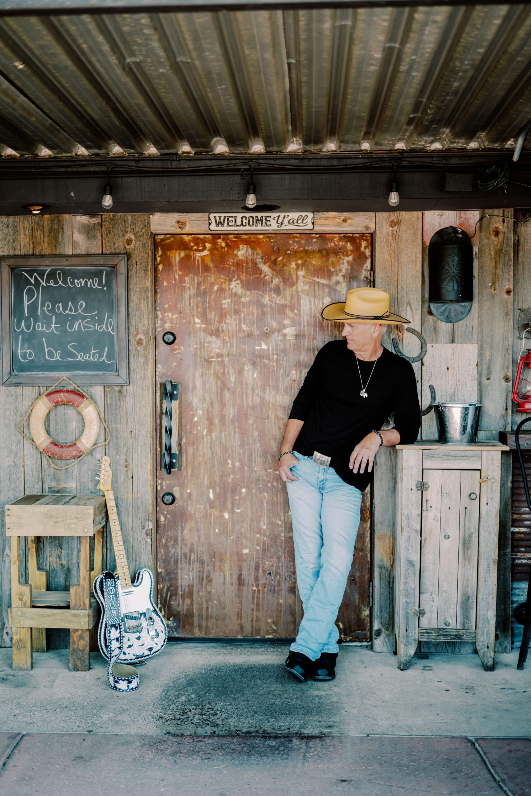 A man leans on a rustic porch in a black shirt, hat and jeans while staring at his guitar during his personal branding session