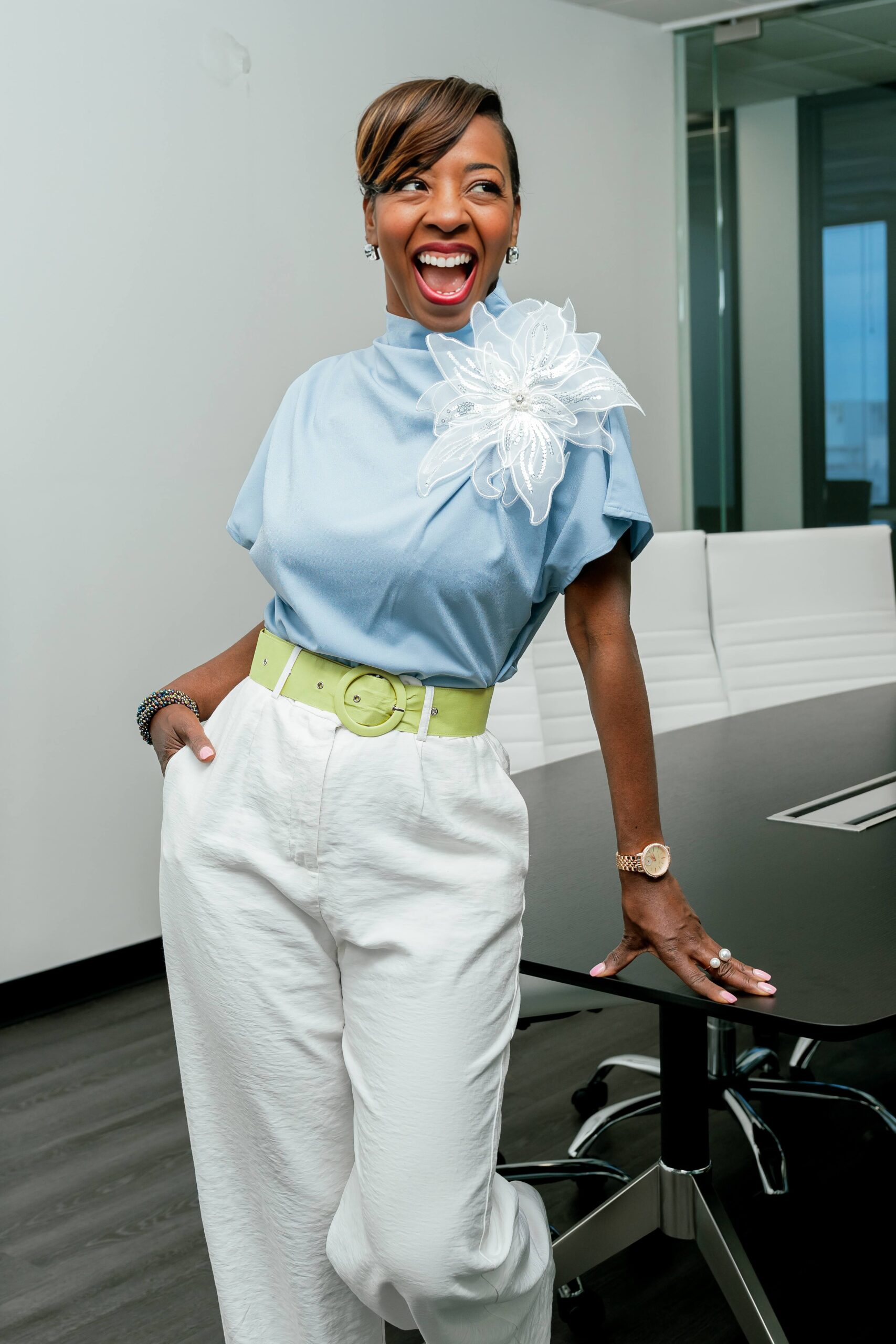 A woman smiles big while leaning on a desk in a blue blouse and white pants during branding photos after getting professional hair and makeup