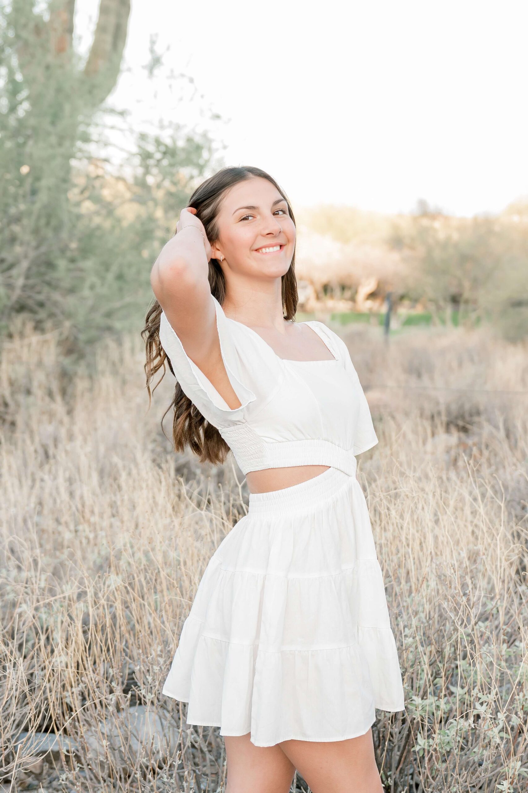 A smiling woman in a white dress pulls her hair back while walking in a park