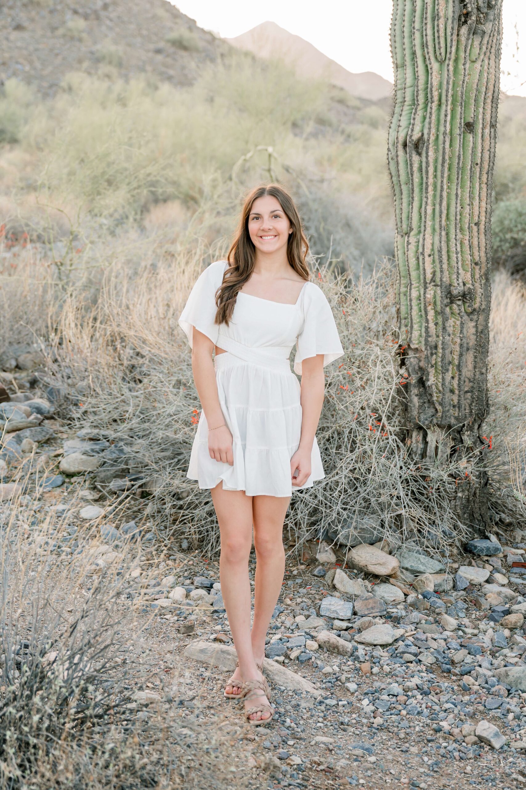 A high school senior smiles while walking a desert trail in a white dress at sunset after finding prom dresses in scottsdale