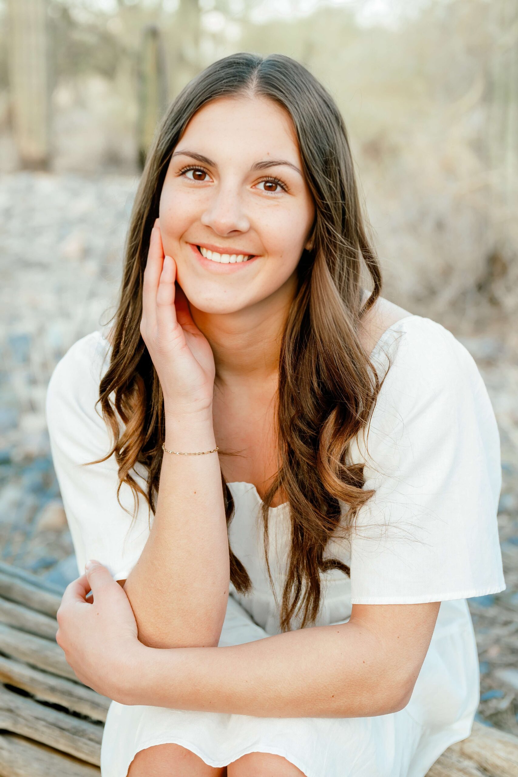 A high school senior in a white dress sits on a log in a desert with a hand on her cheek after finding prom dresses in scottsdale