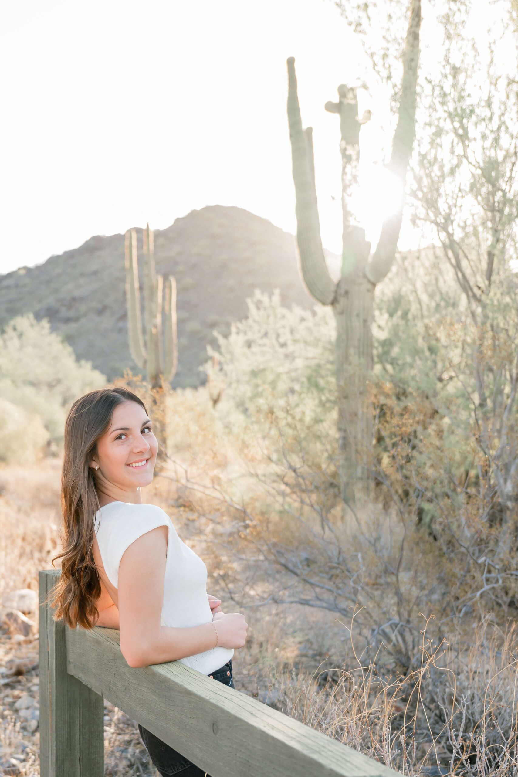 A smiling high school senior in a white blouse leans back on a wooden fence in the desert at sunset
