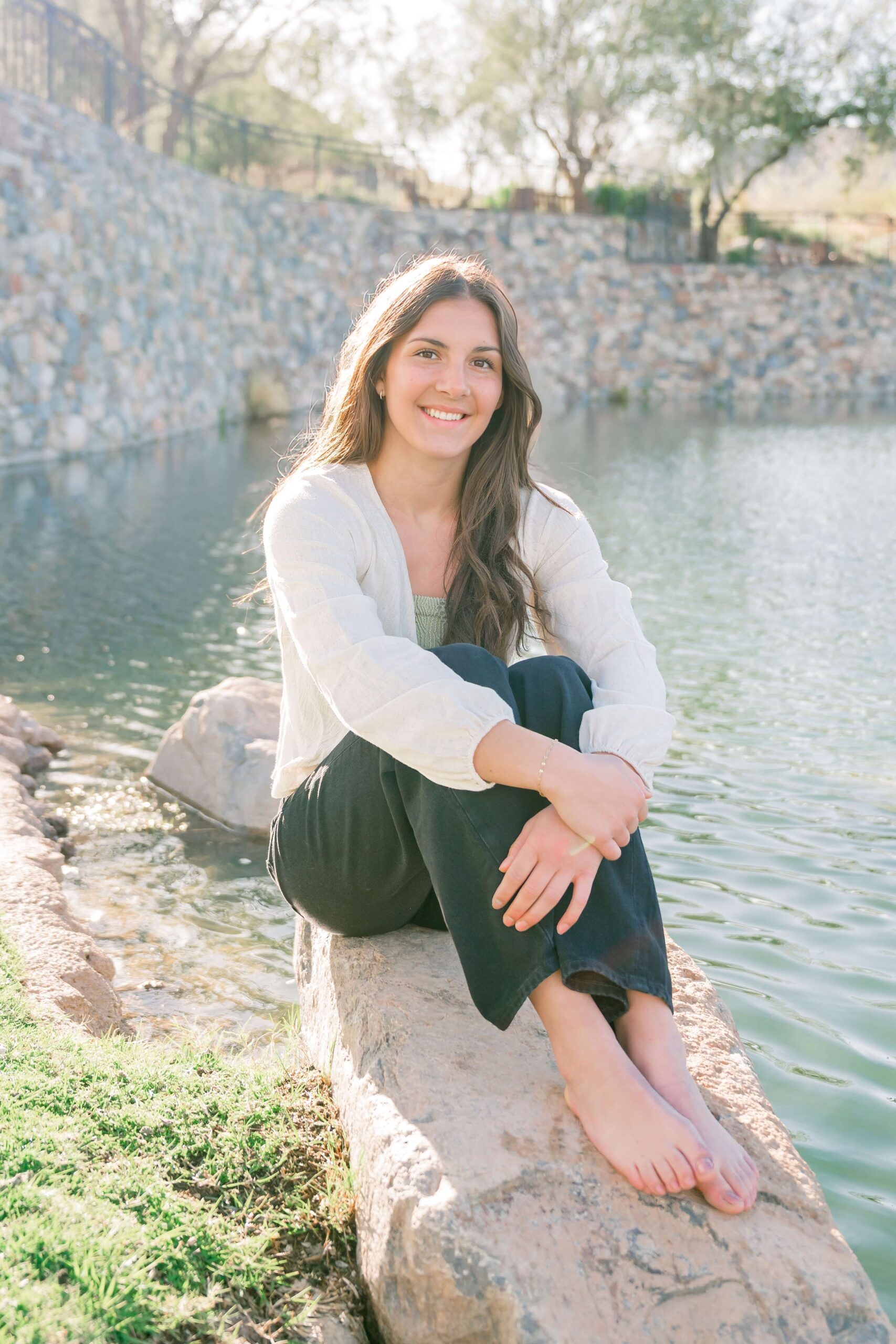 a high school senior sits on the edge of a pond on a boulder after exploring tanning salons in scottsdale
