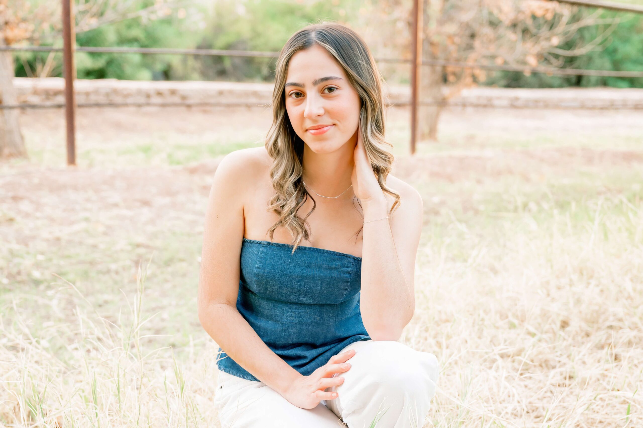 A high school senior in a denim blouse kneels in a field of tall grass after finding tanning salons in scottsdale
