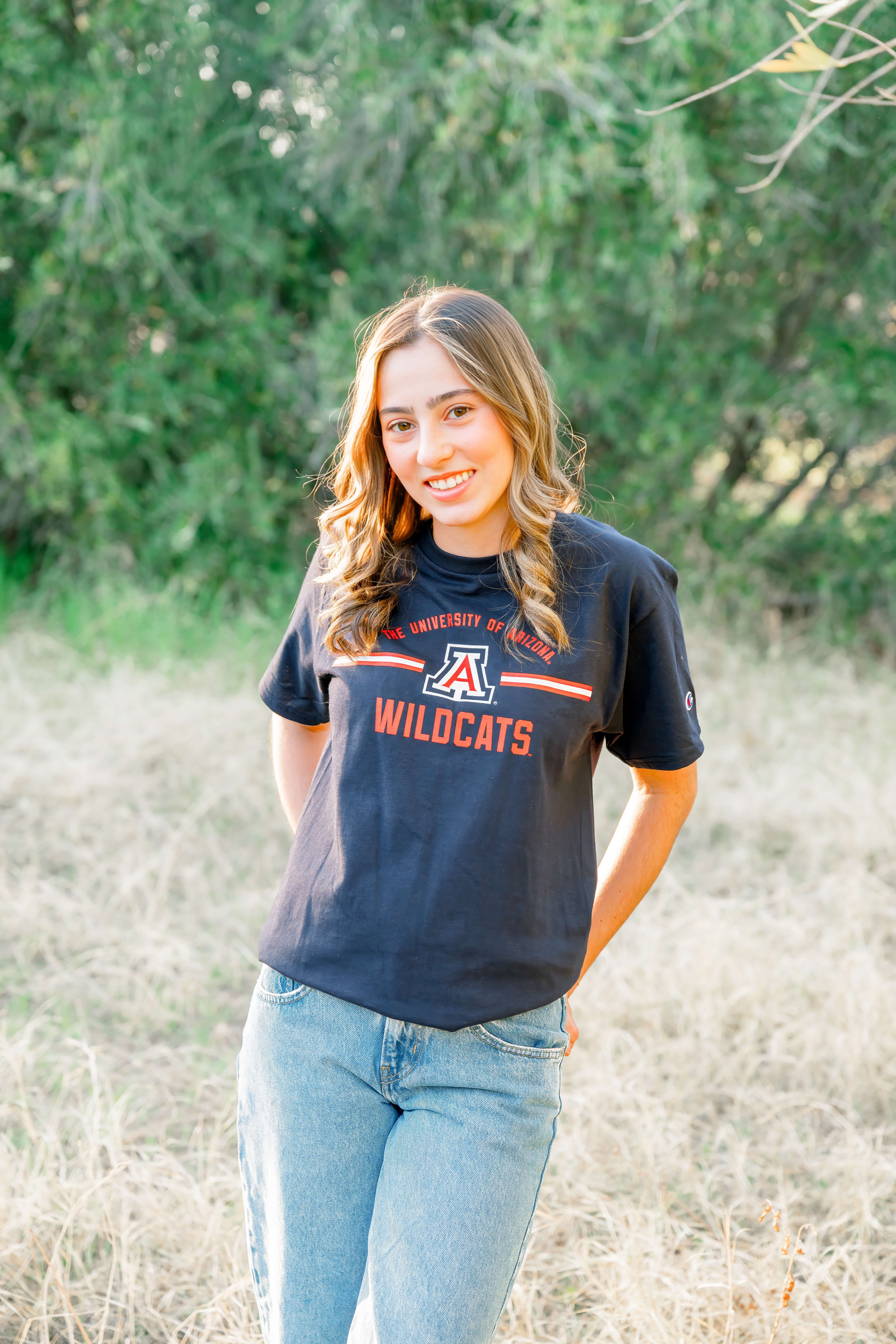 A high school senior stands in a college shirt with hands in her pockets at sunset