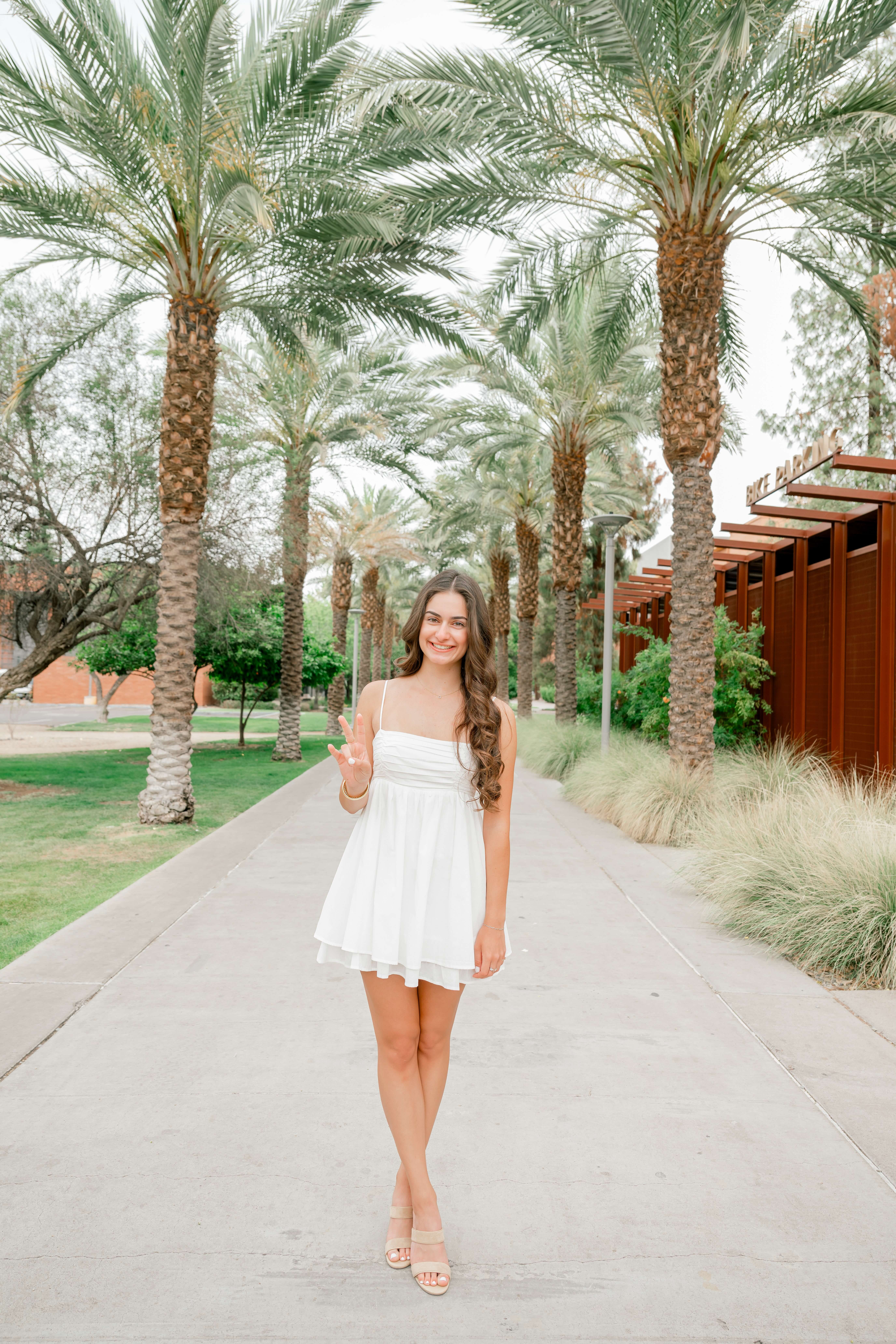 A high school senior in a white dress gives a peace sign in a sidewalk lined with palms after shopping for prom dresses in phoenix