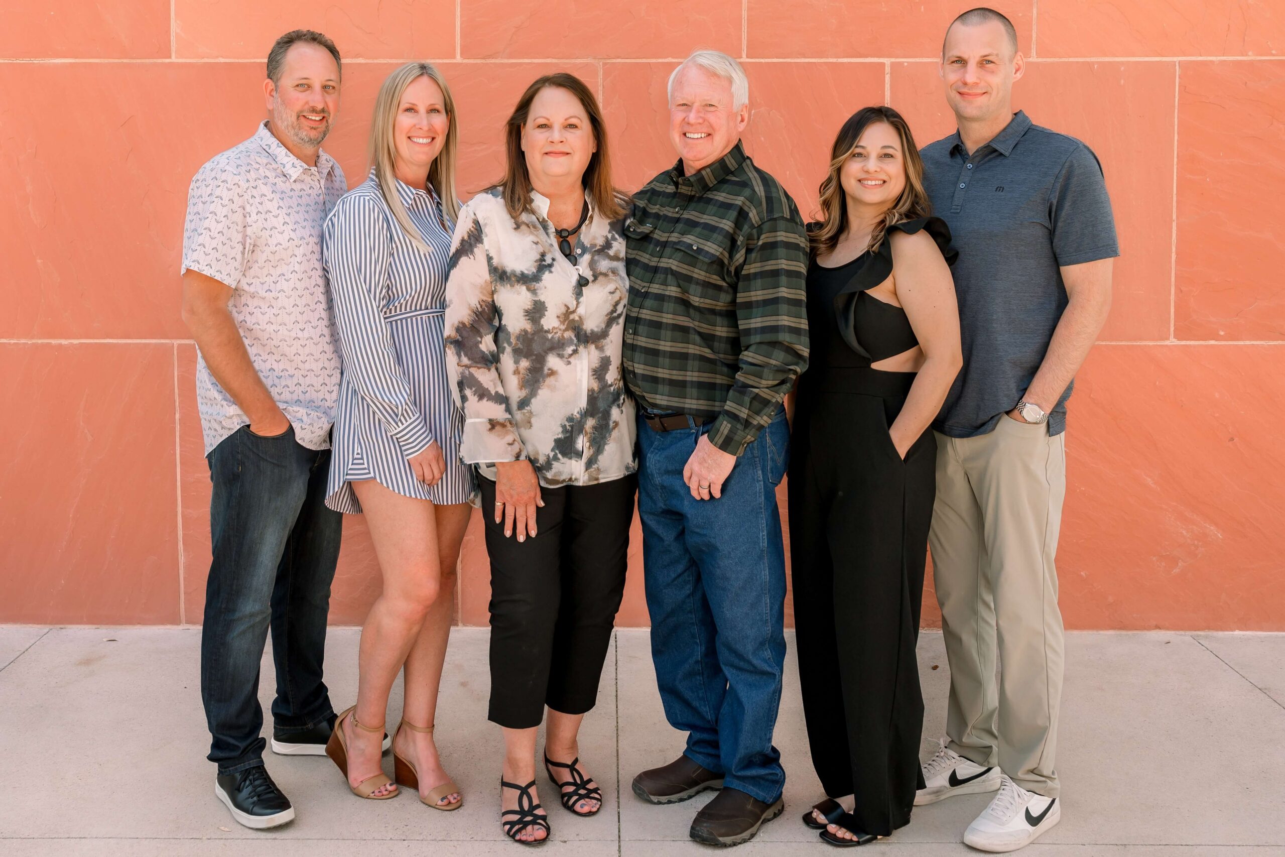 An extended family stand smiling against an orange wall with hands in pockets