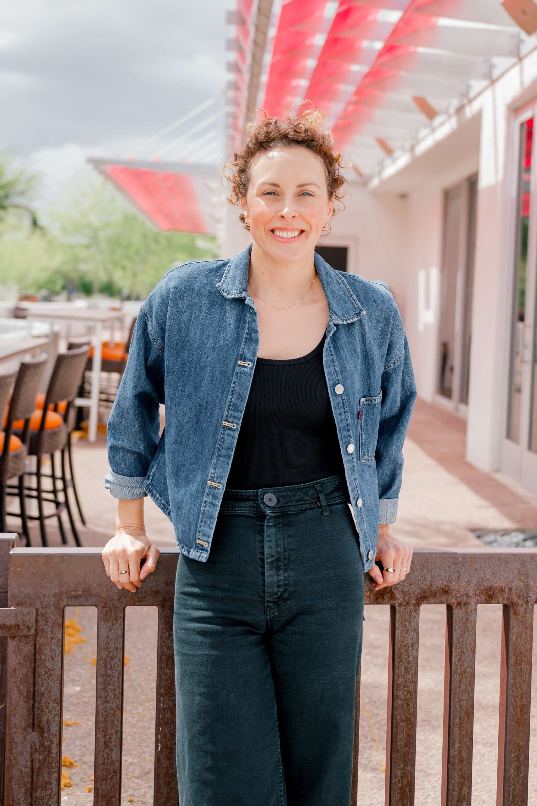 A woman smiles while leaning on a patio riling in a denim jacket for her personal branding photos