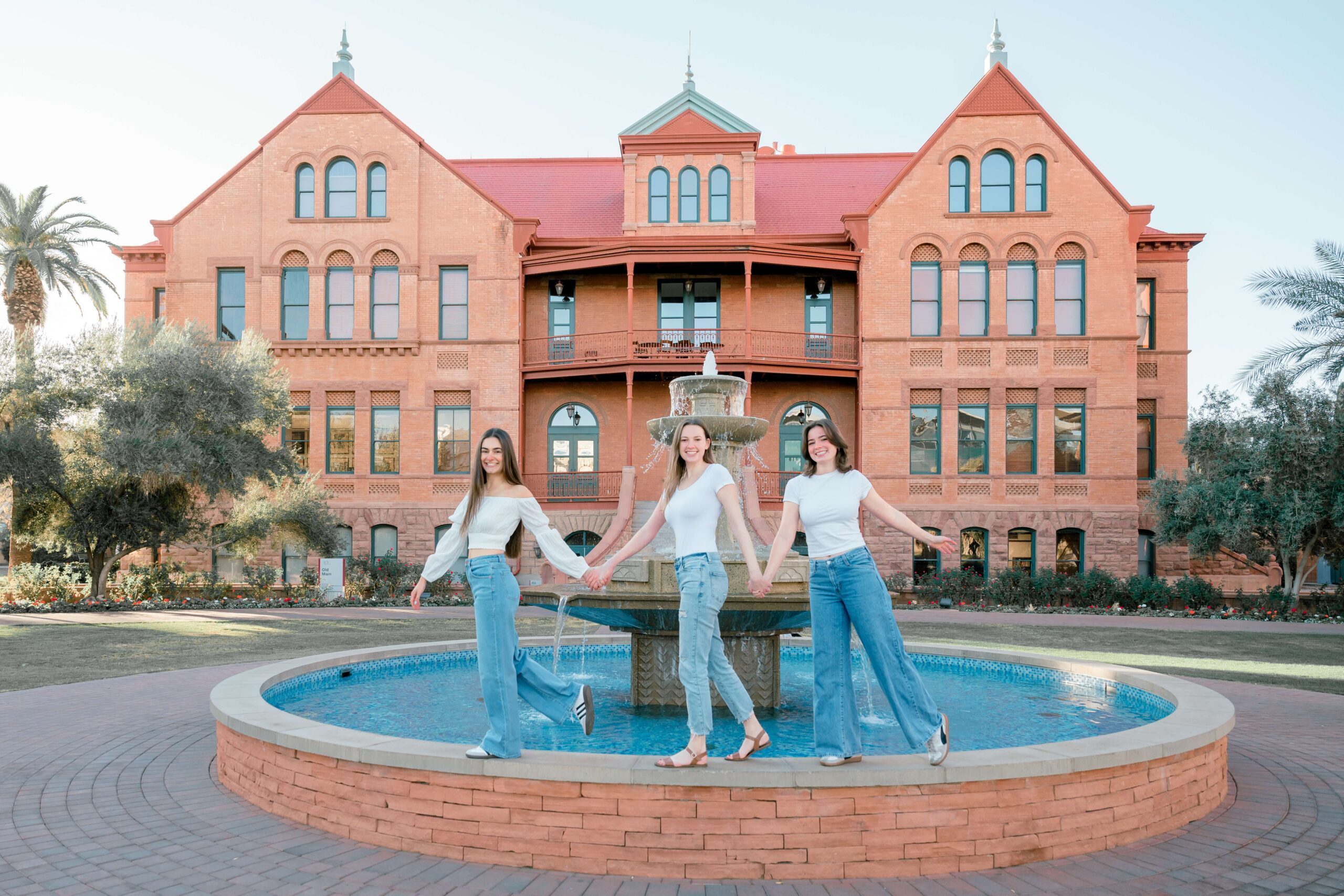 Three college grads walk holding hands on a fountain ledge in matching white shirts and jeans