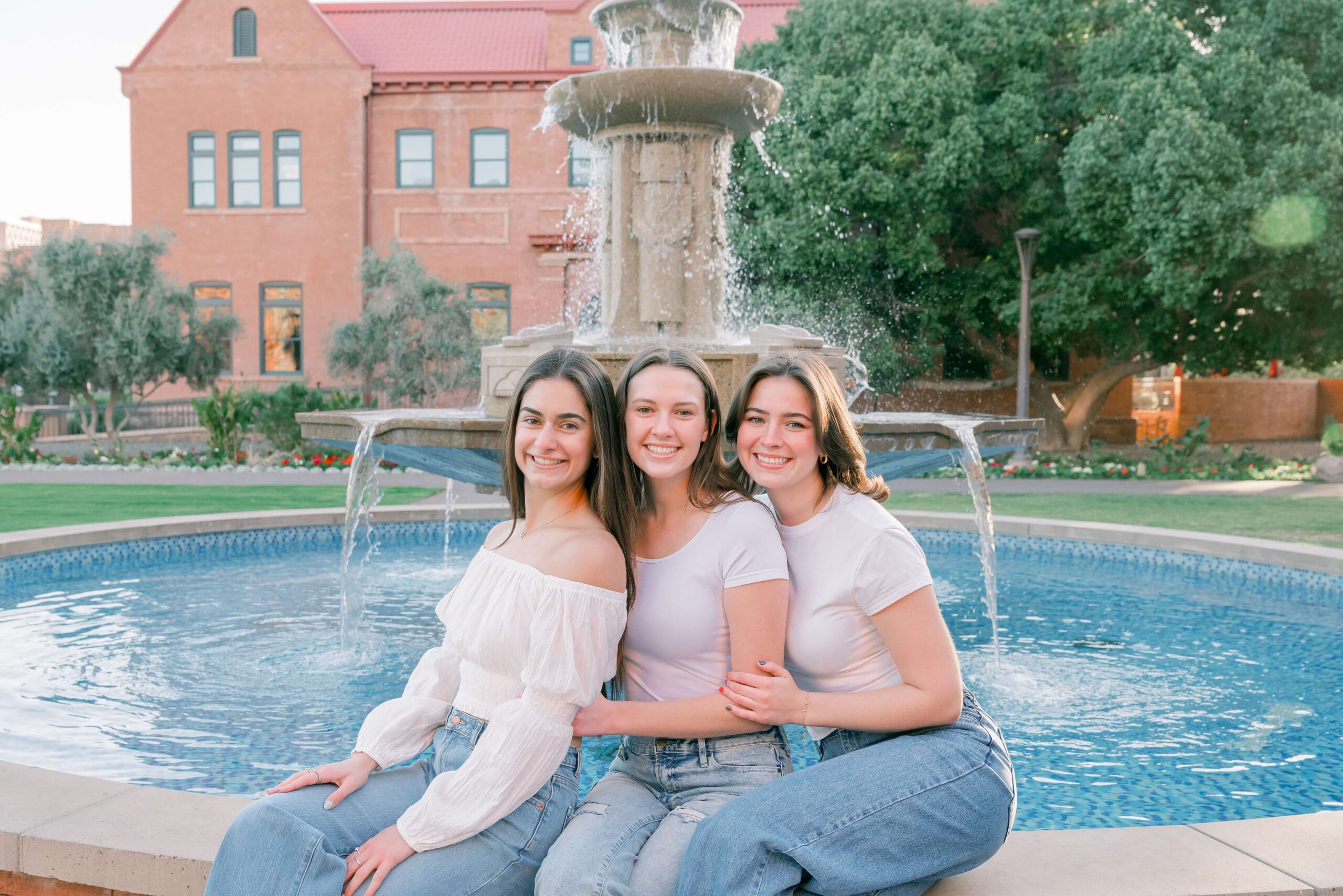 Three best friends in matching white and jeans sit smiling together on a fountain ledge after finding tanning salons in phoenix