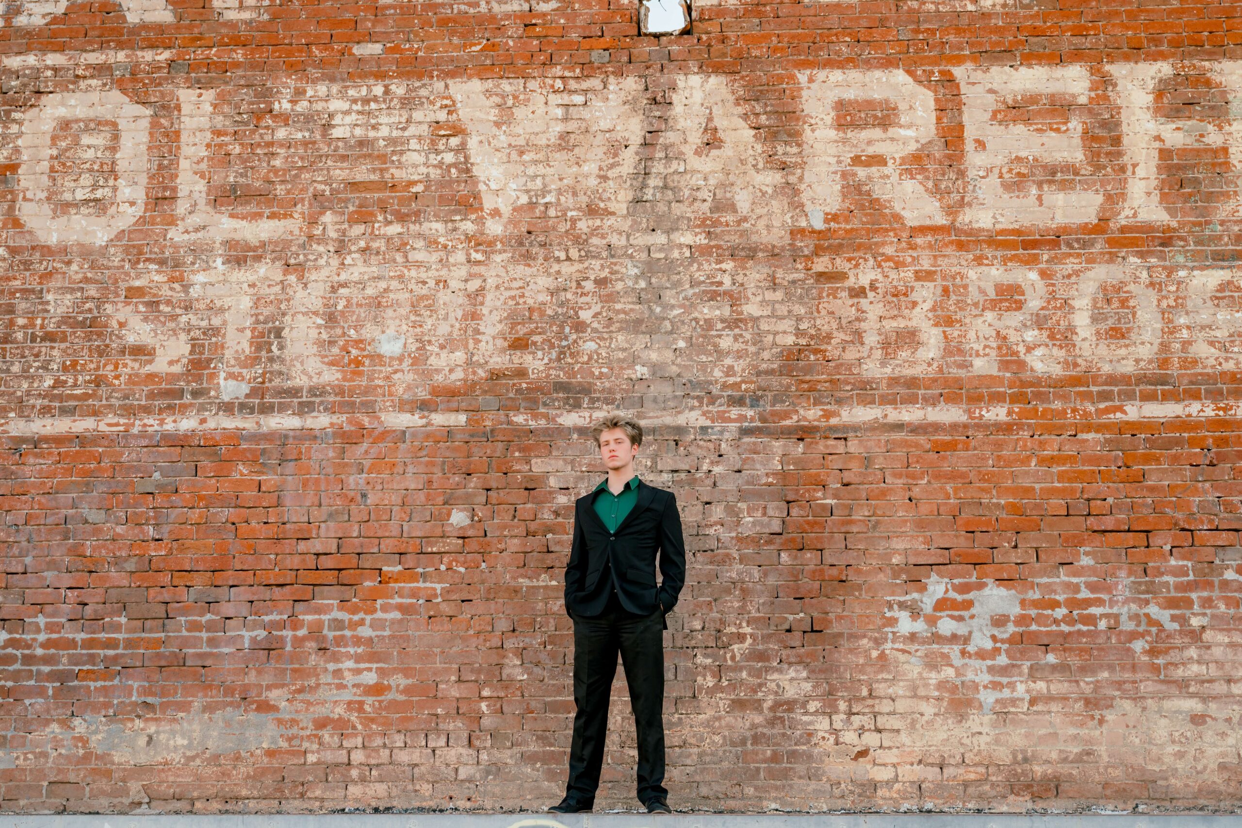 A high school grad stands with his hands in his pockets against a rustic brick wall with old paint