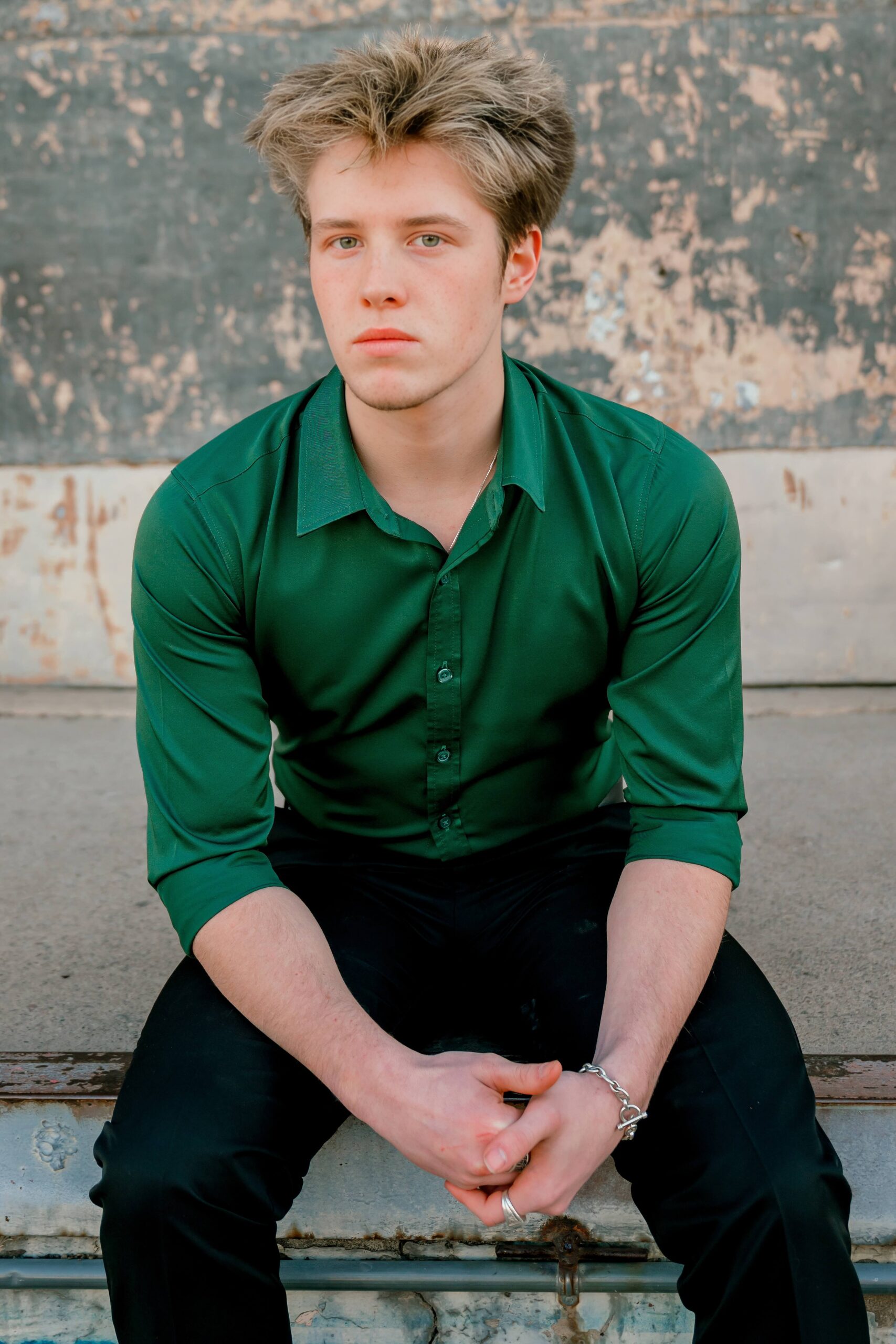 A high school senior in a green button down sits on a loading dock leaning on his knees after finding tux rental in phoenix