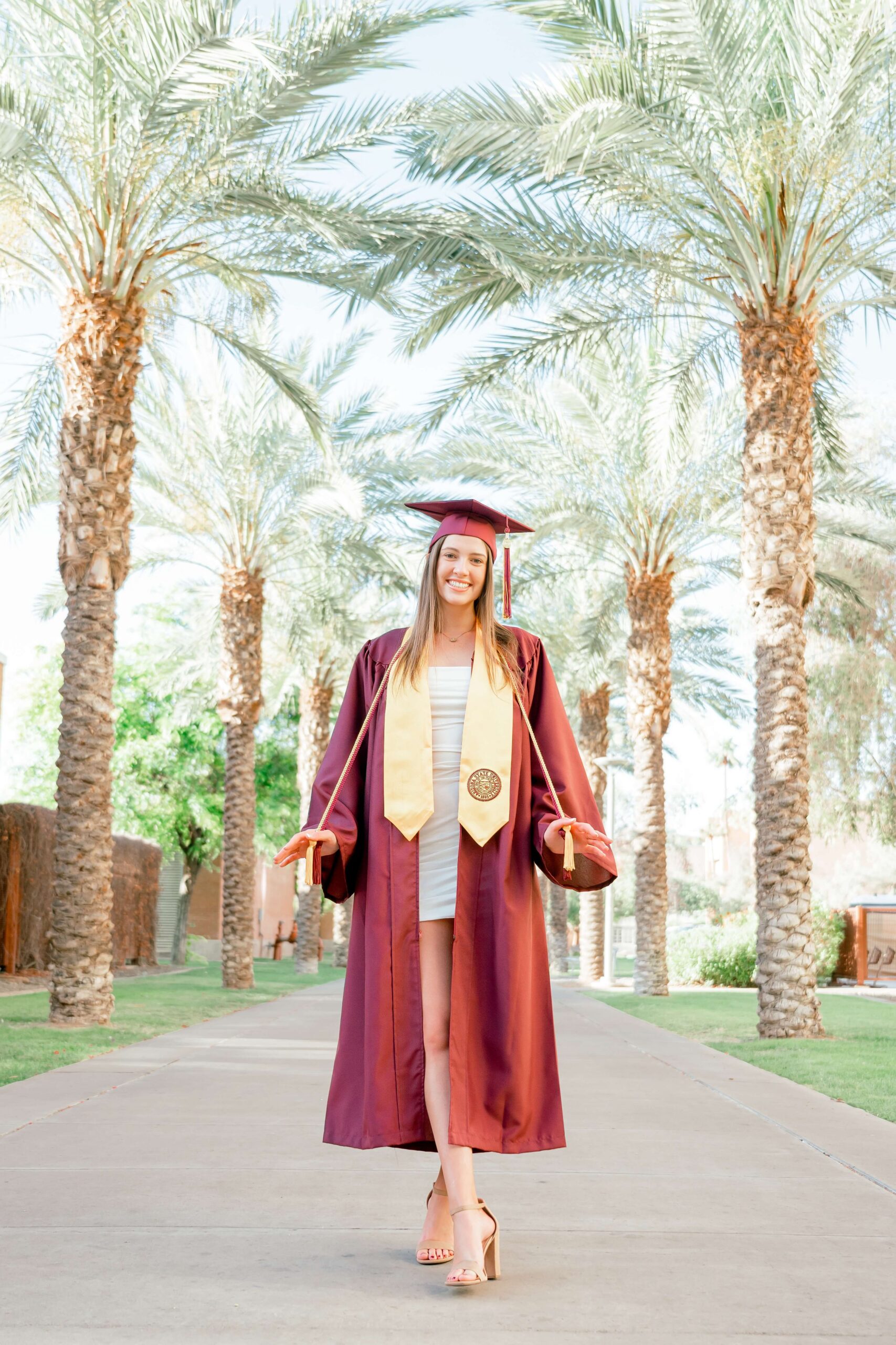 A happy grad stands in a palm lined sidewalk in her cap and gown holding chords and wearing a gold stole