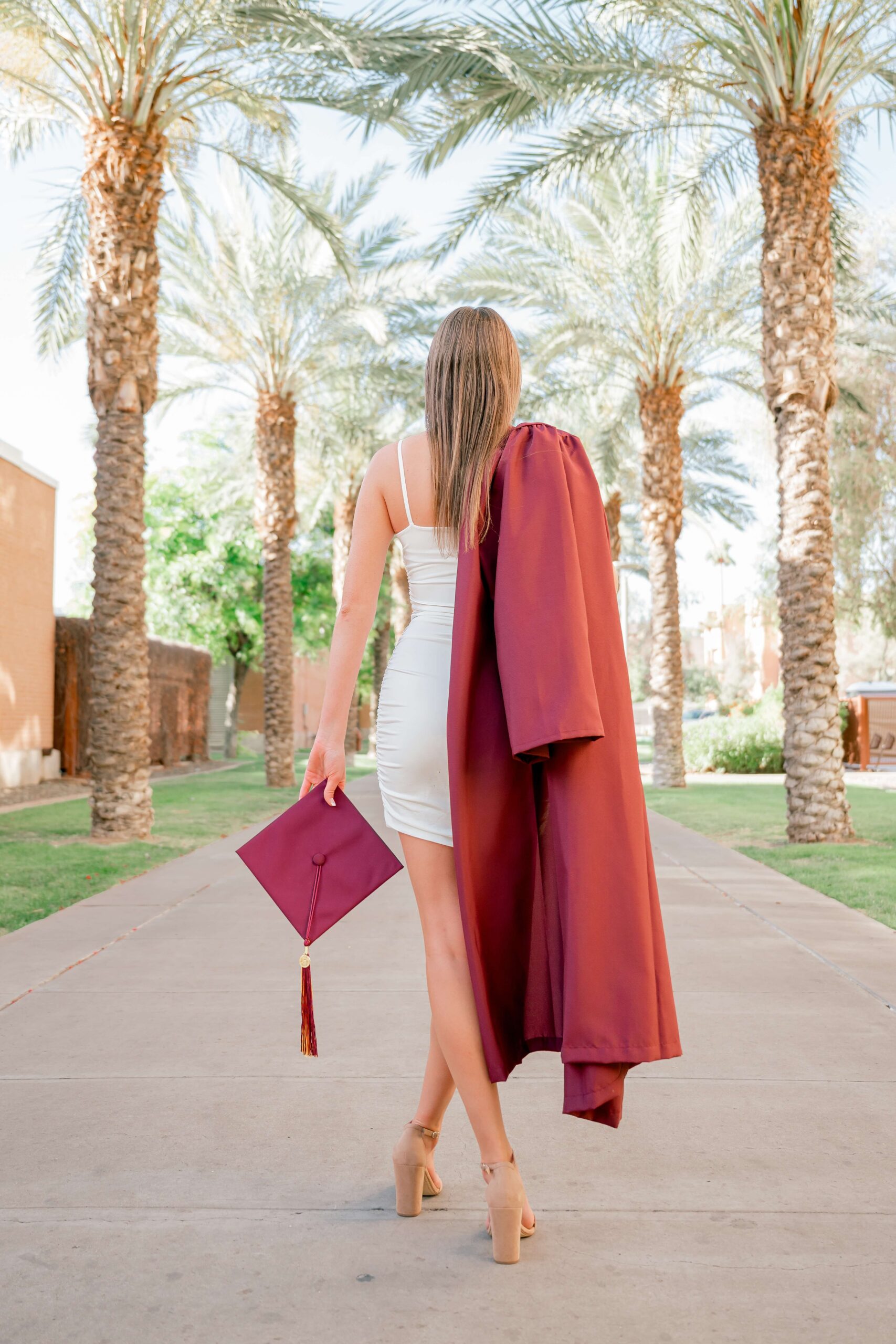 A graduate walks with her gown over her shoulder and cap in hand in a white dress on a palm lined sidewalk at one of the Colleges near phoenix