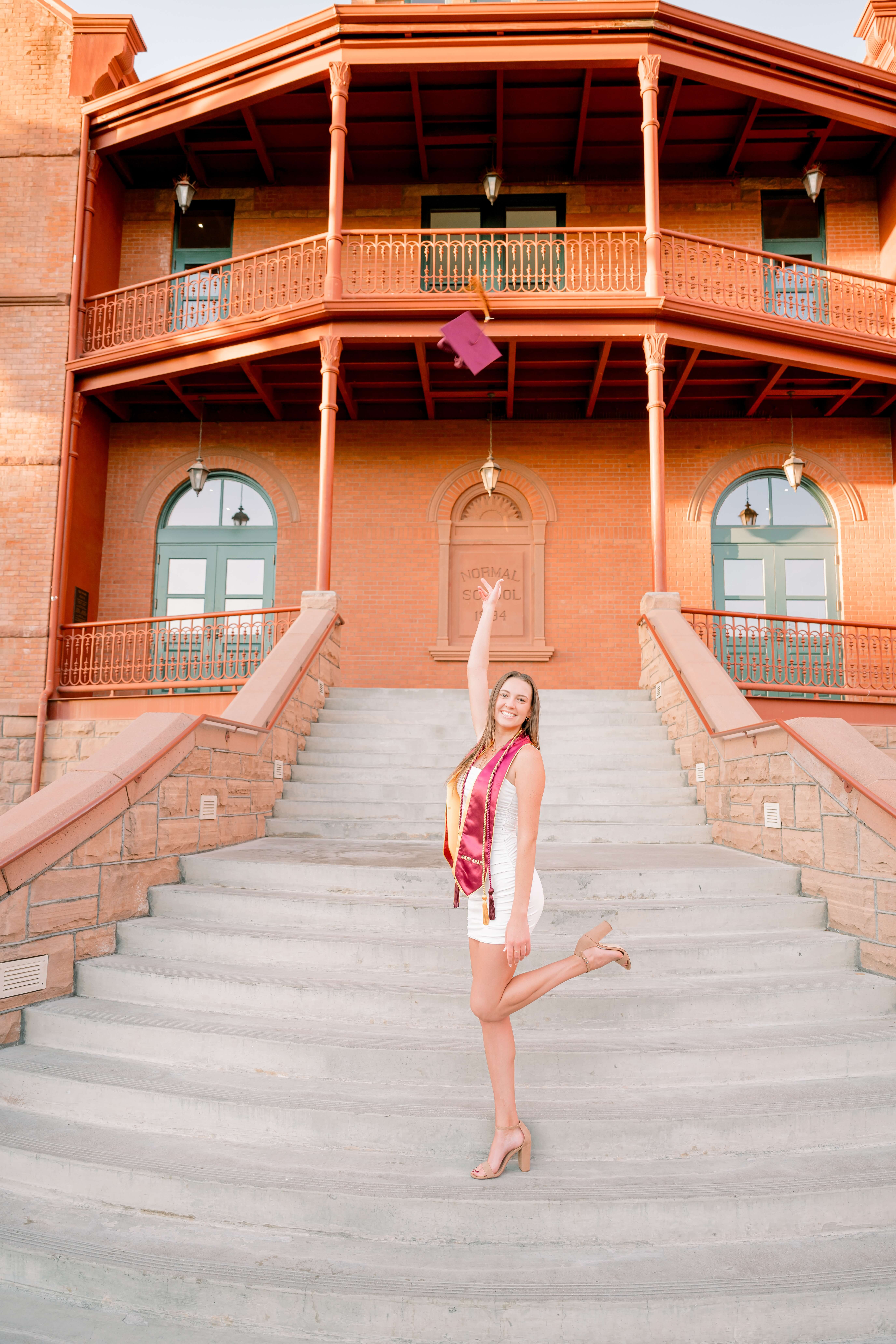 A graduate in a white dress and red stole tosses her cap in the air on the steps of an orange building at one of the Colleges near phoenix