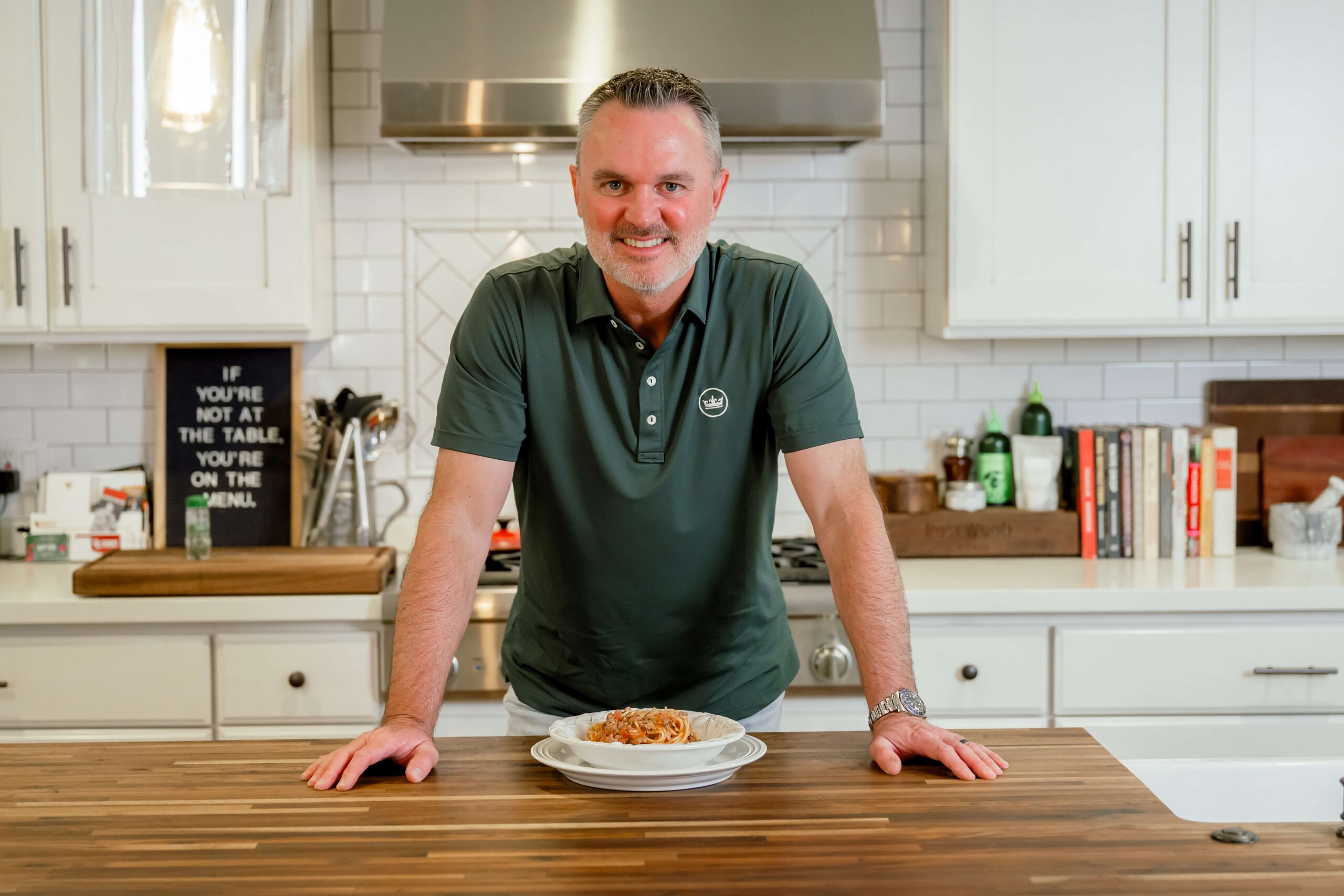 A man smiles while leaning on a kitchen island over a bowl of spaghetti