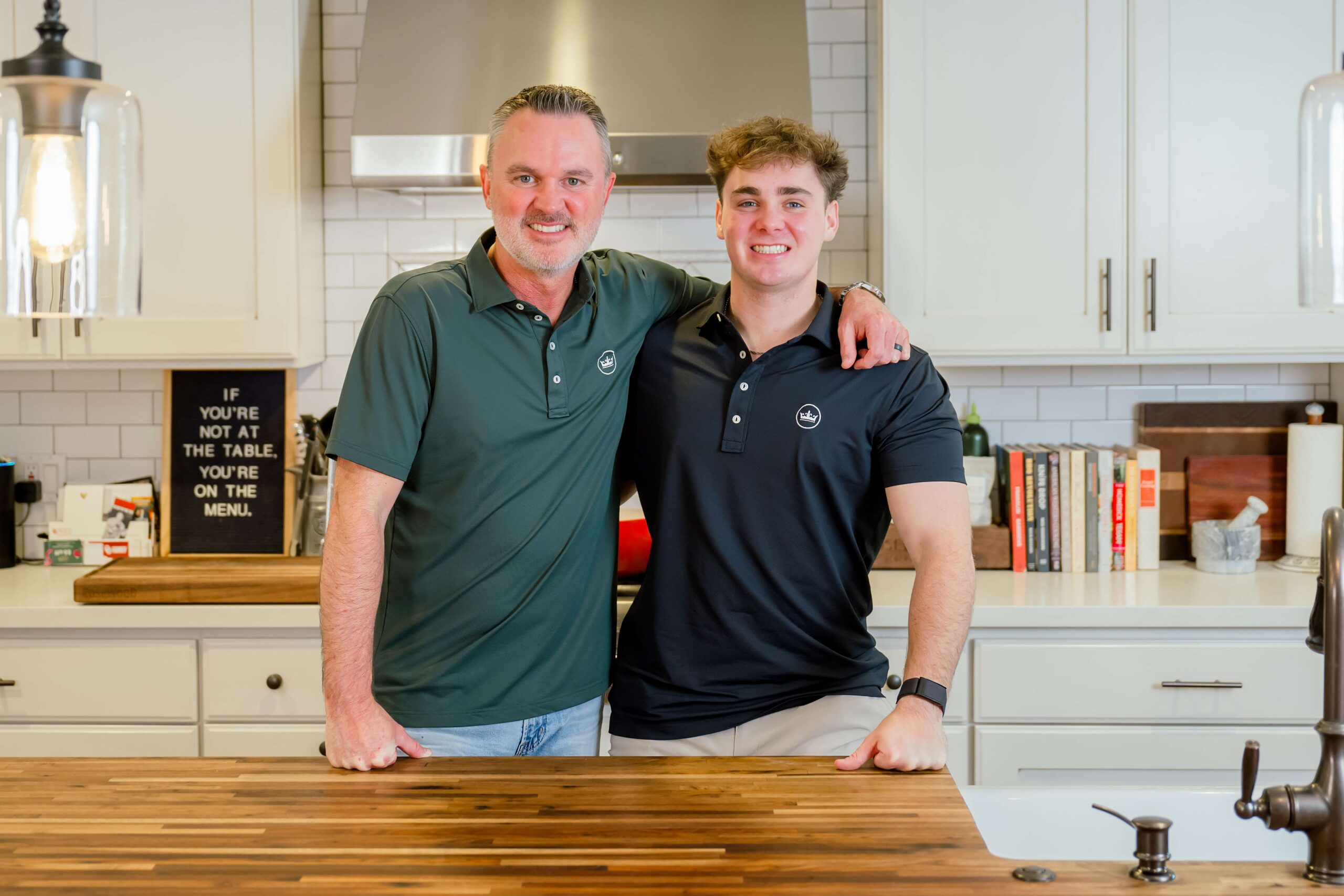 A dad in a green polo stands in a kitchen stands in the kitchen with his arm around his son in a matching black polo