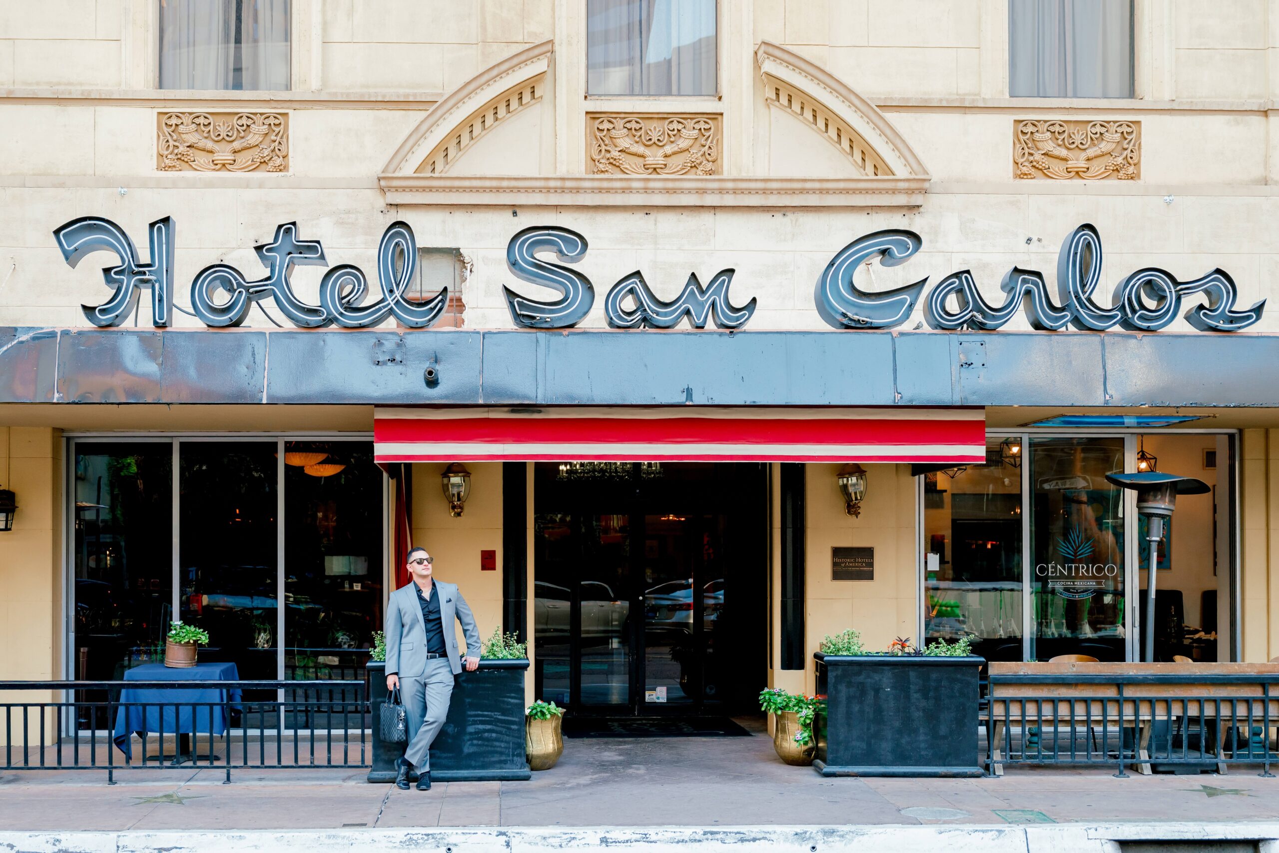 A man leans on a planter in front of a hotel restaurant in a grey suit and sunglasses