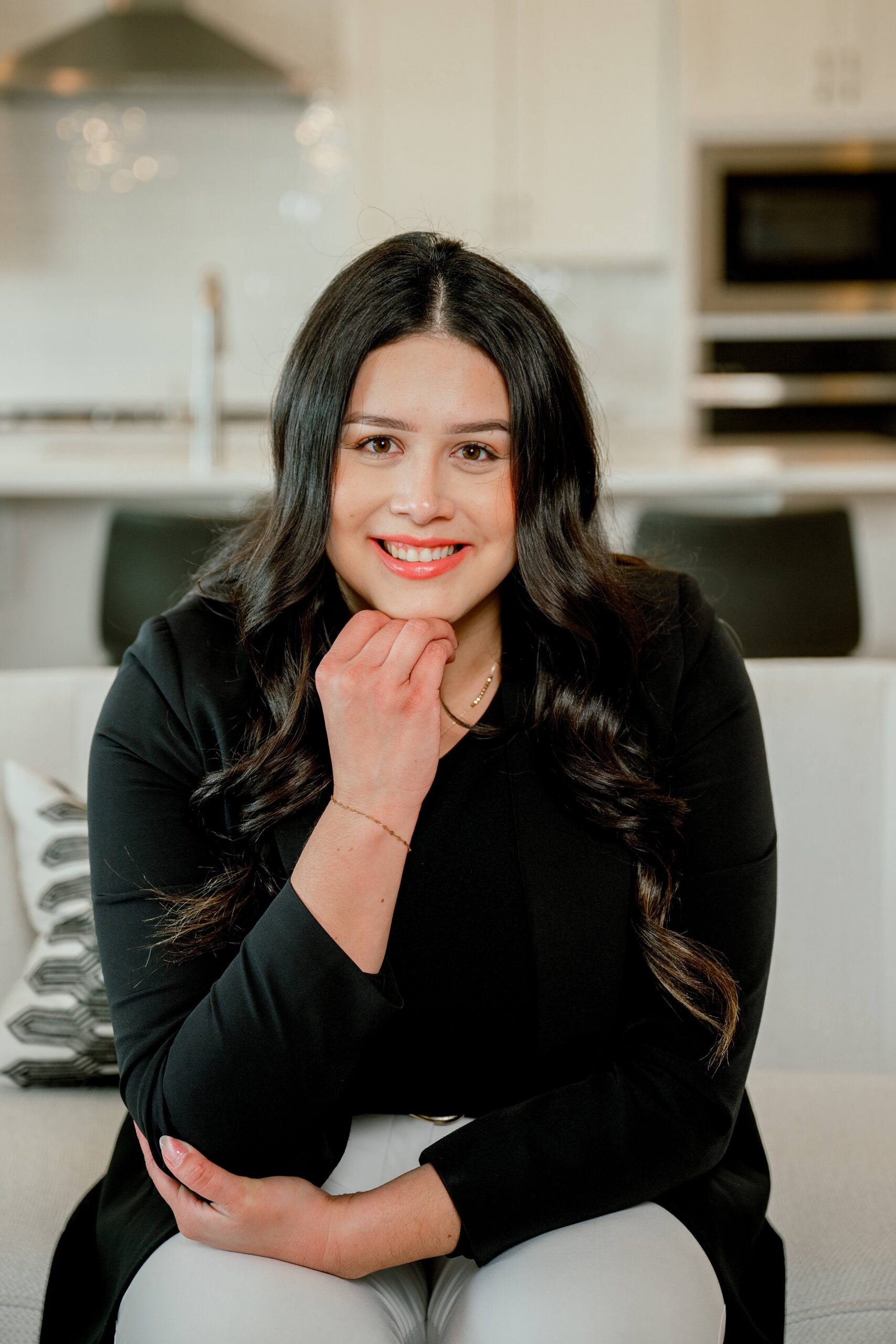 A woman leans on her hand and knee while sitting on a couch in a living room in a black blouse