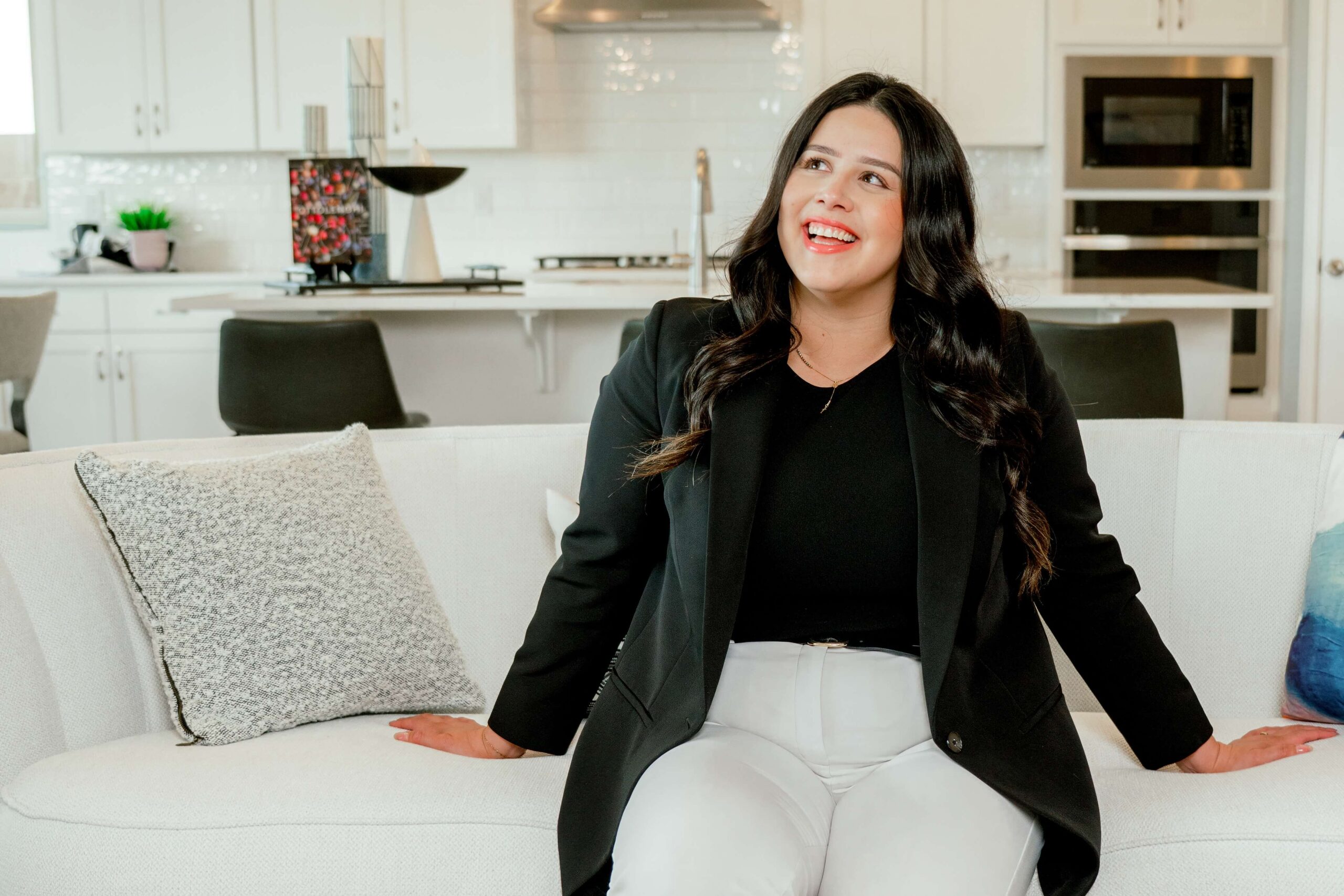 A happy woman smiles while sitting on a couch in a black blazer and capturing must-have brand photos