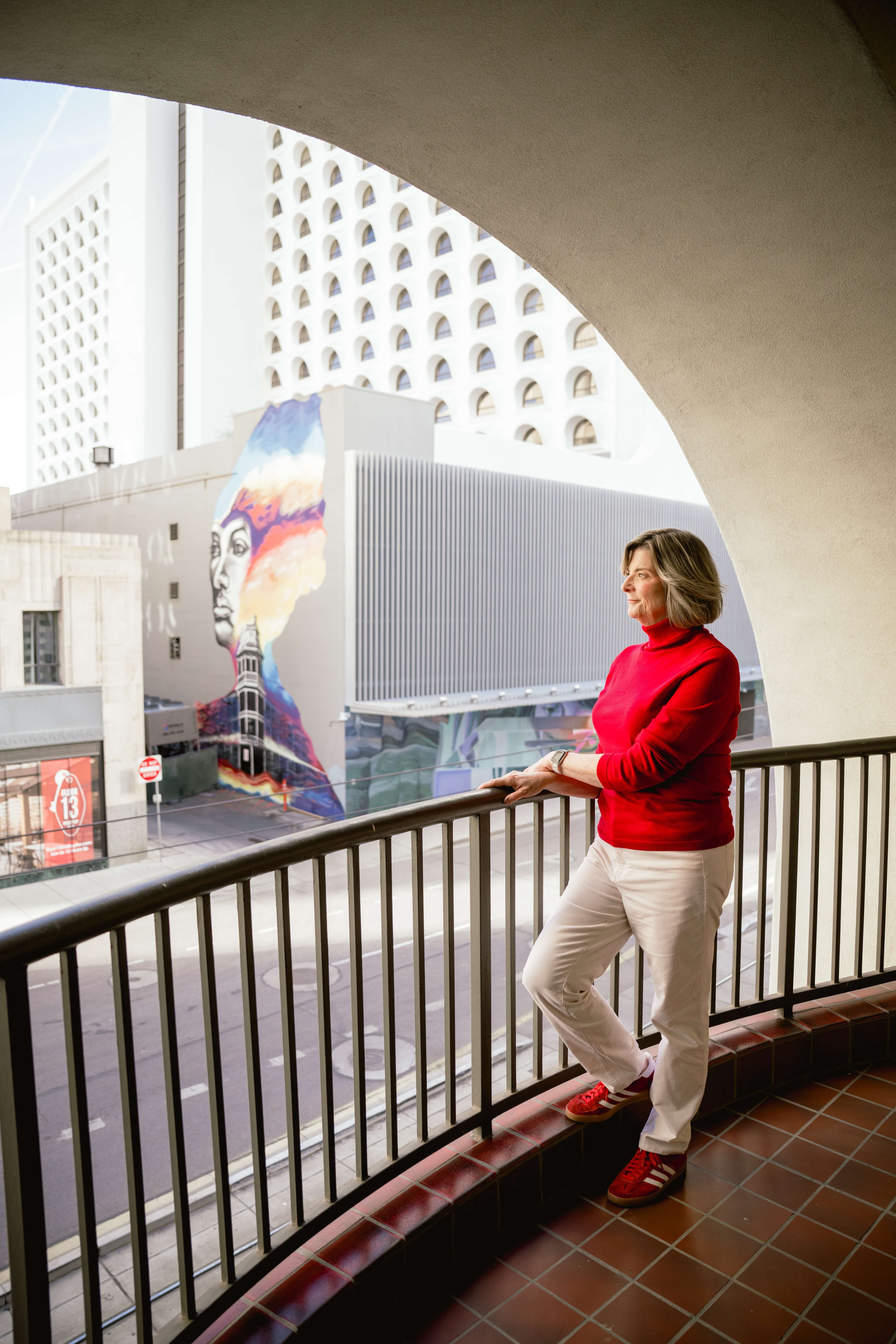 A woman in a red sweater stands against a balcony gazing out to the sunset in downtown