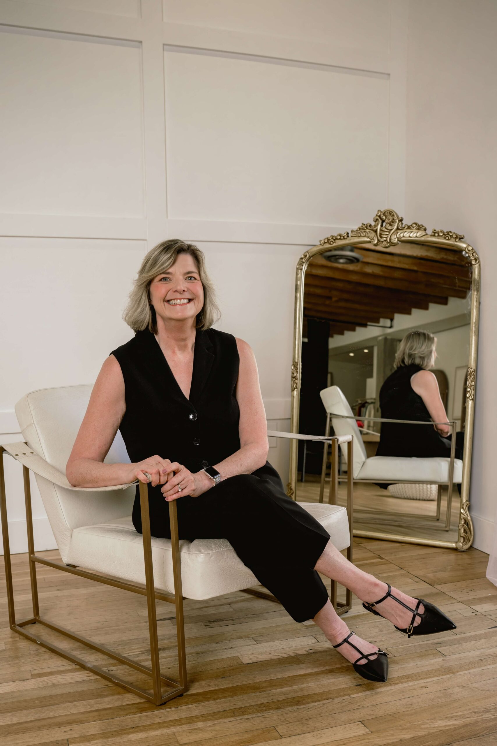 A woman sits in a chair in front of a mirror smiling in a black outfit