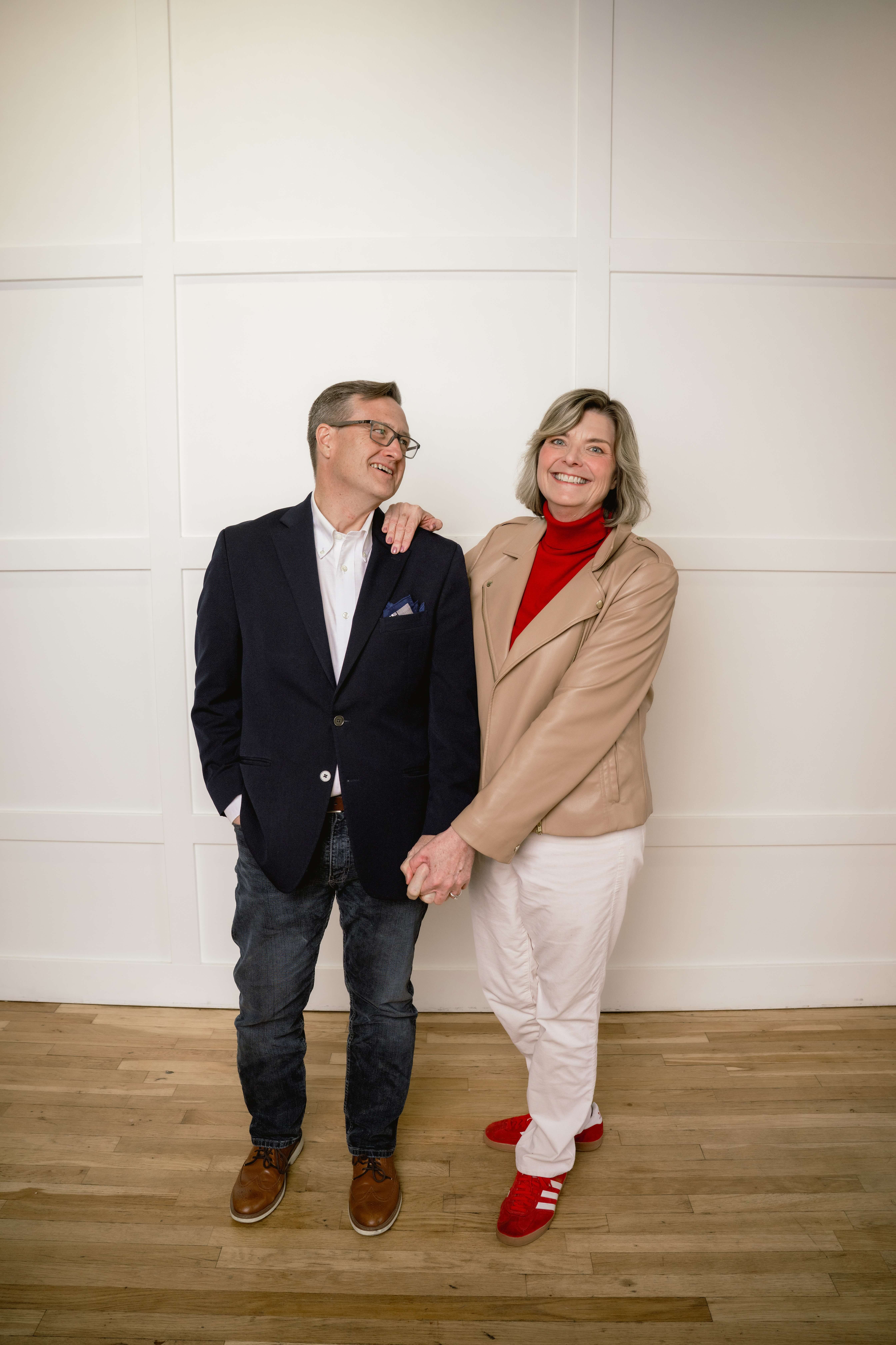 A happy couple in black and tan stand against a wall holding hands and smiling while posing for brand photos