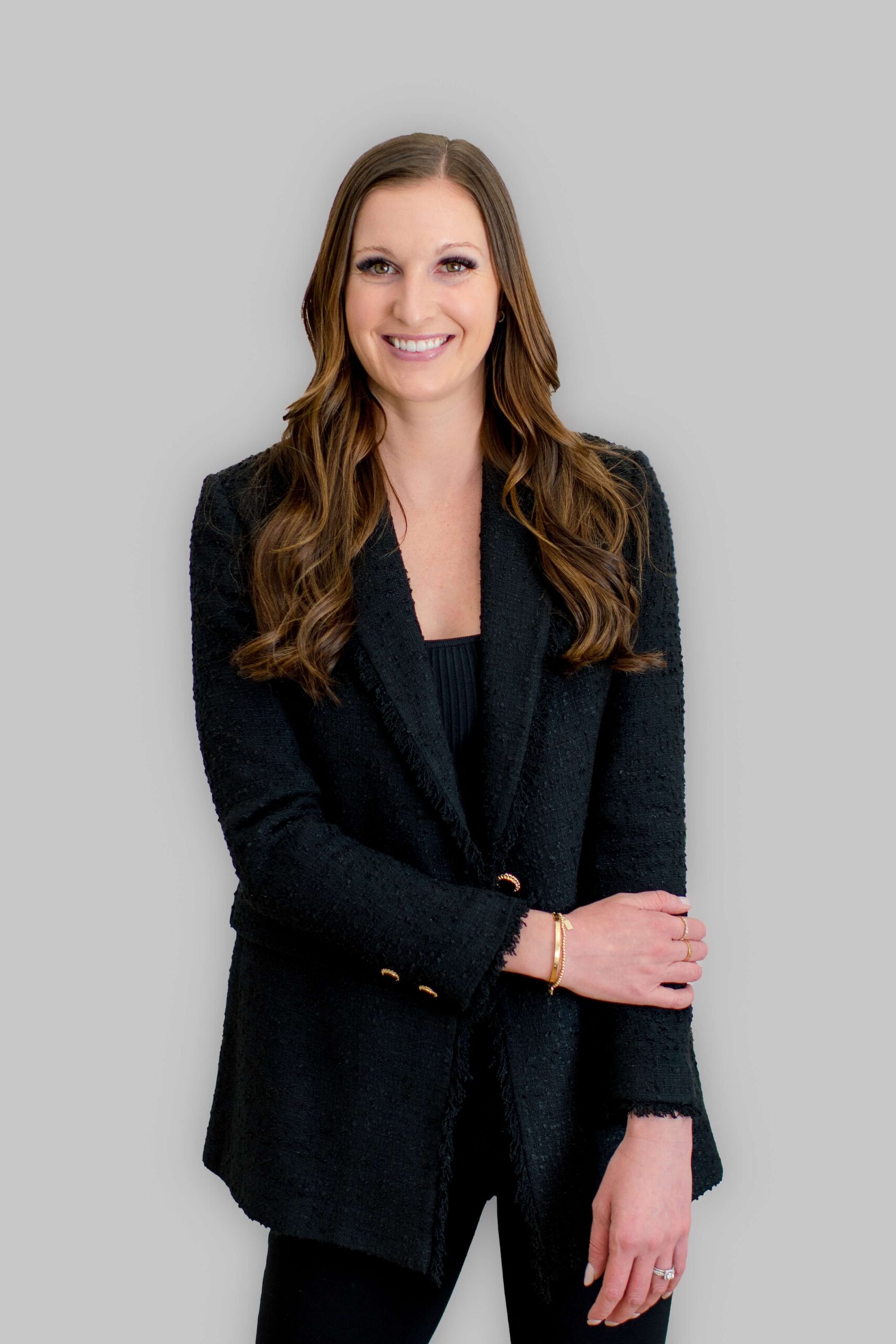 A happy woman stands in a black jacket against a white wall with a smile