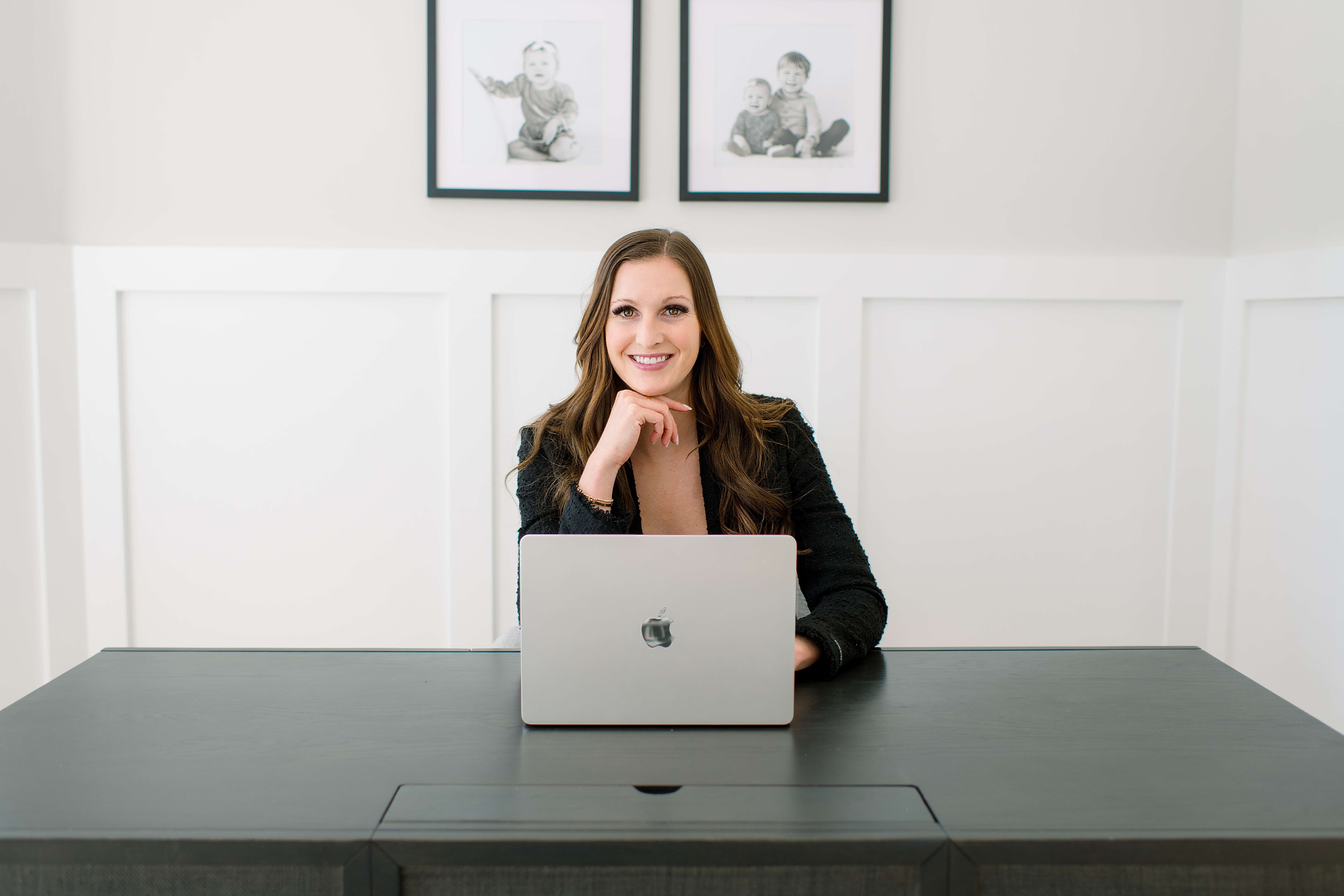 A woman in a black jacket sits behind a desk smiling with a laptop during her realtor branding photos
